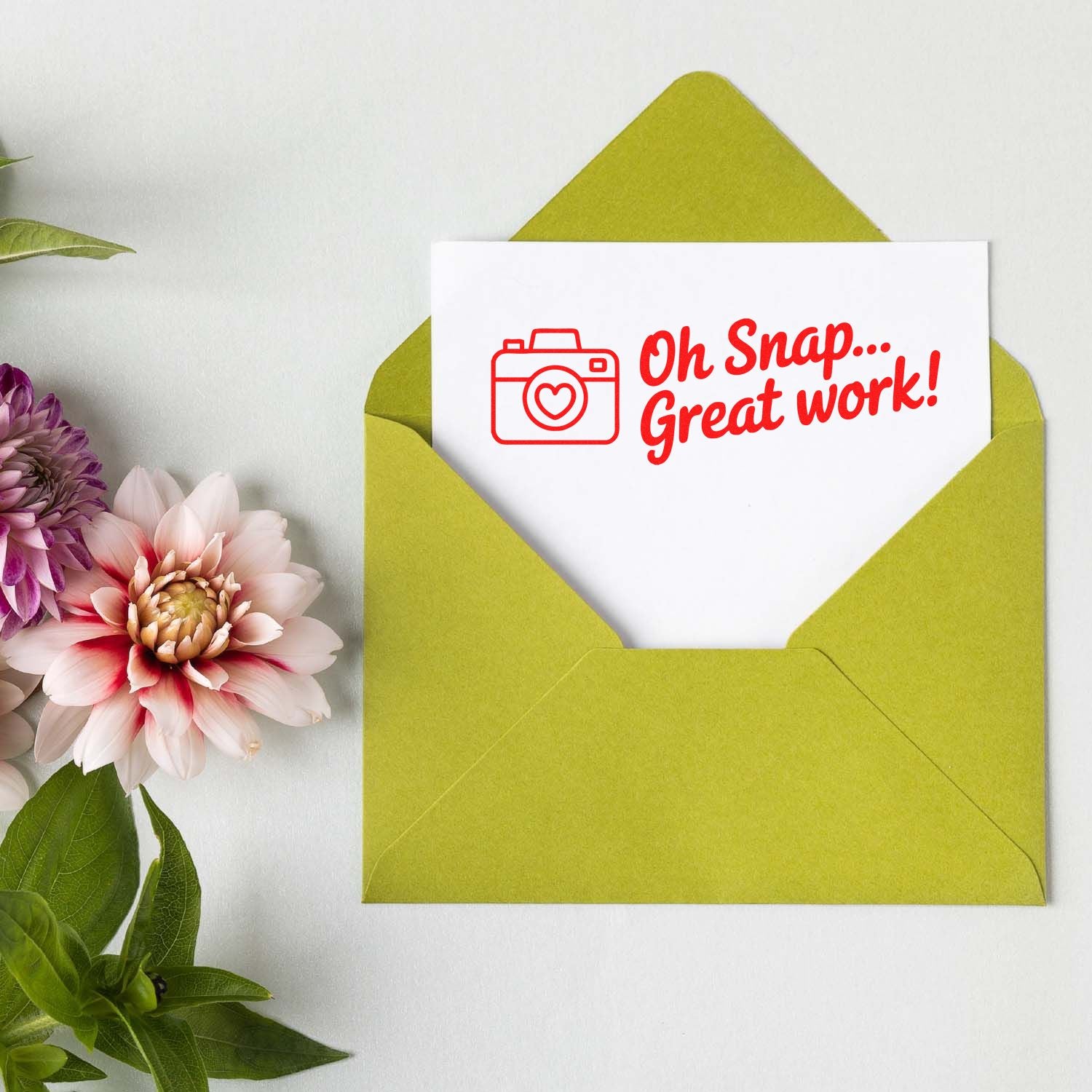 Green envelope with a white card featuring a red stamp seal: camera icon and Oh Snap... Great work!, beside pink and purple flowers.