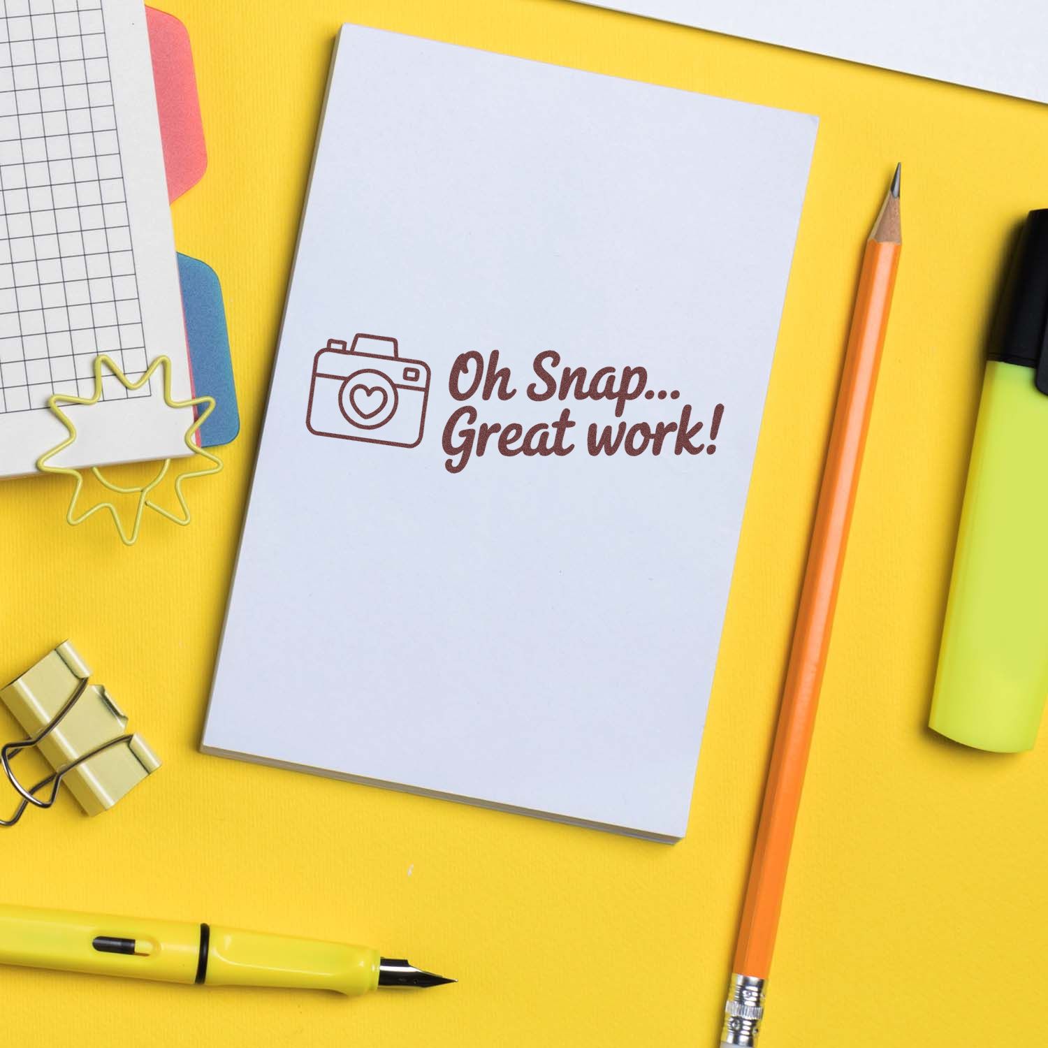White stamp card with brown camera seal reading Oh Snap... Great work! on a bright yellow desk with pencil and stationery.