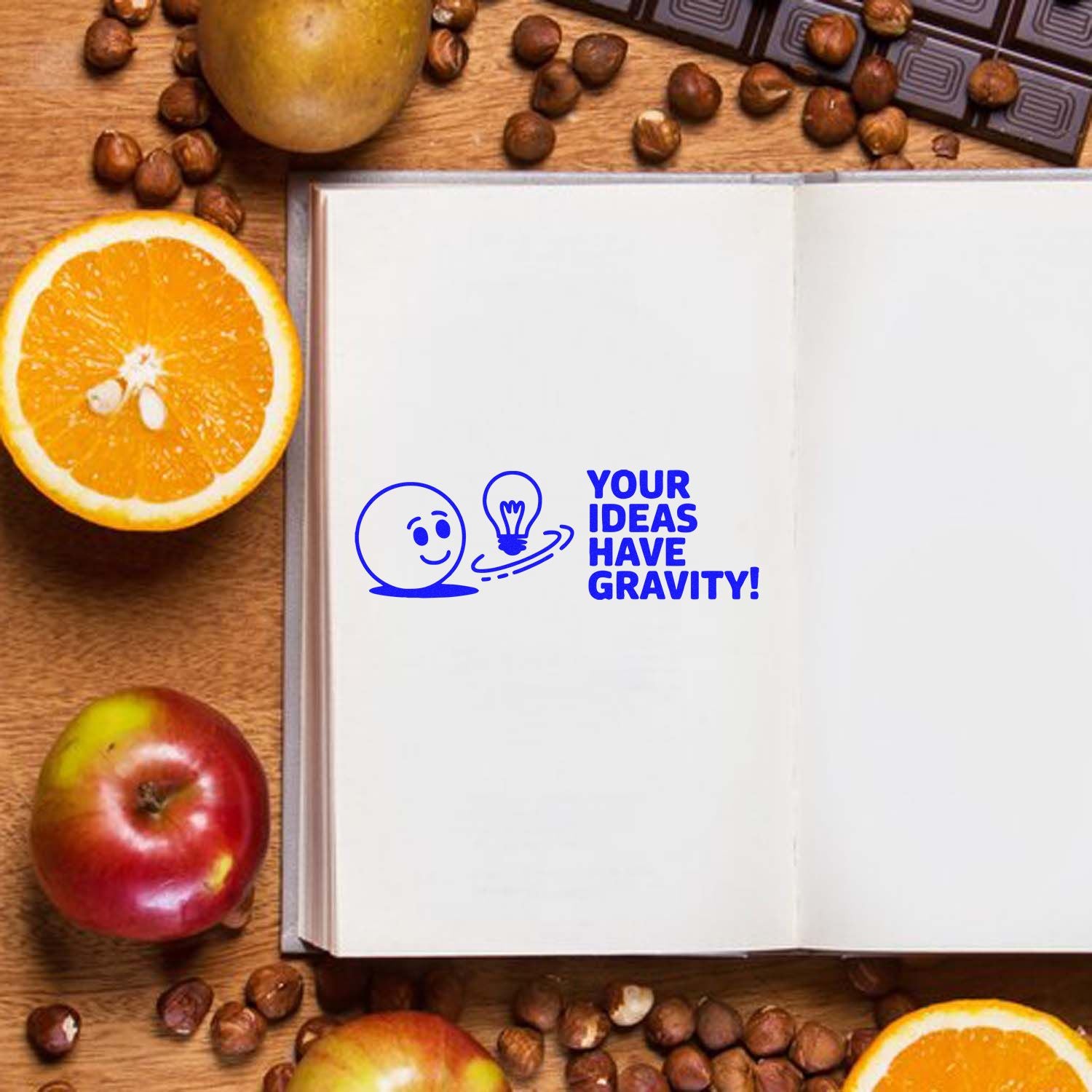 Blue stamp seal on open white notebook, with smiling face, lightbulb and text Your ideas have gravity! surrounded by oranges and apples.