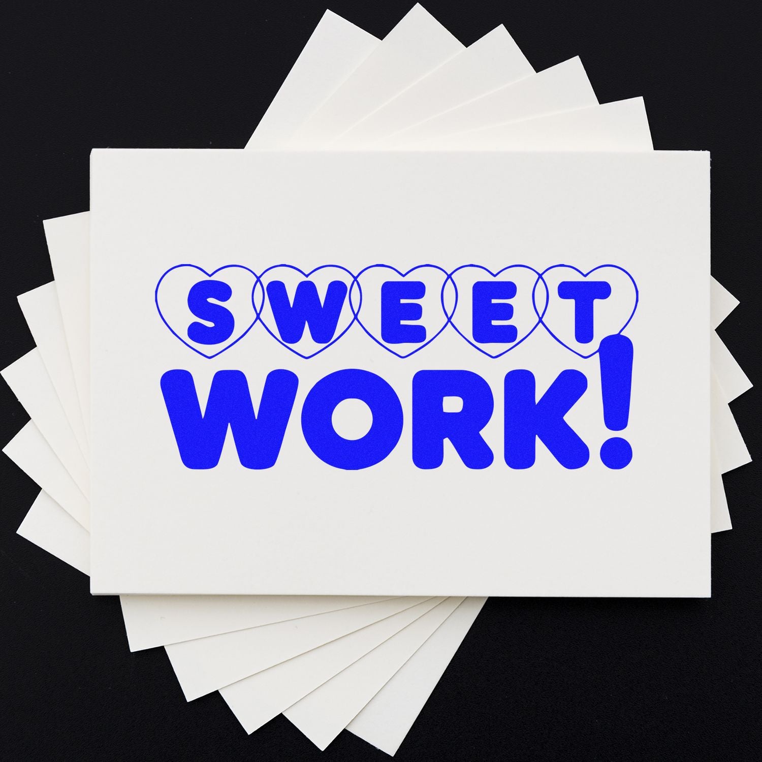 Blue SWEET WORK! stamp with heart outlines on fanned white seal cards against a black background
