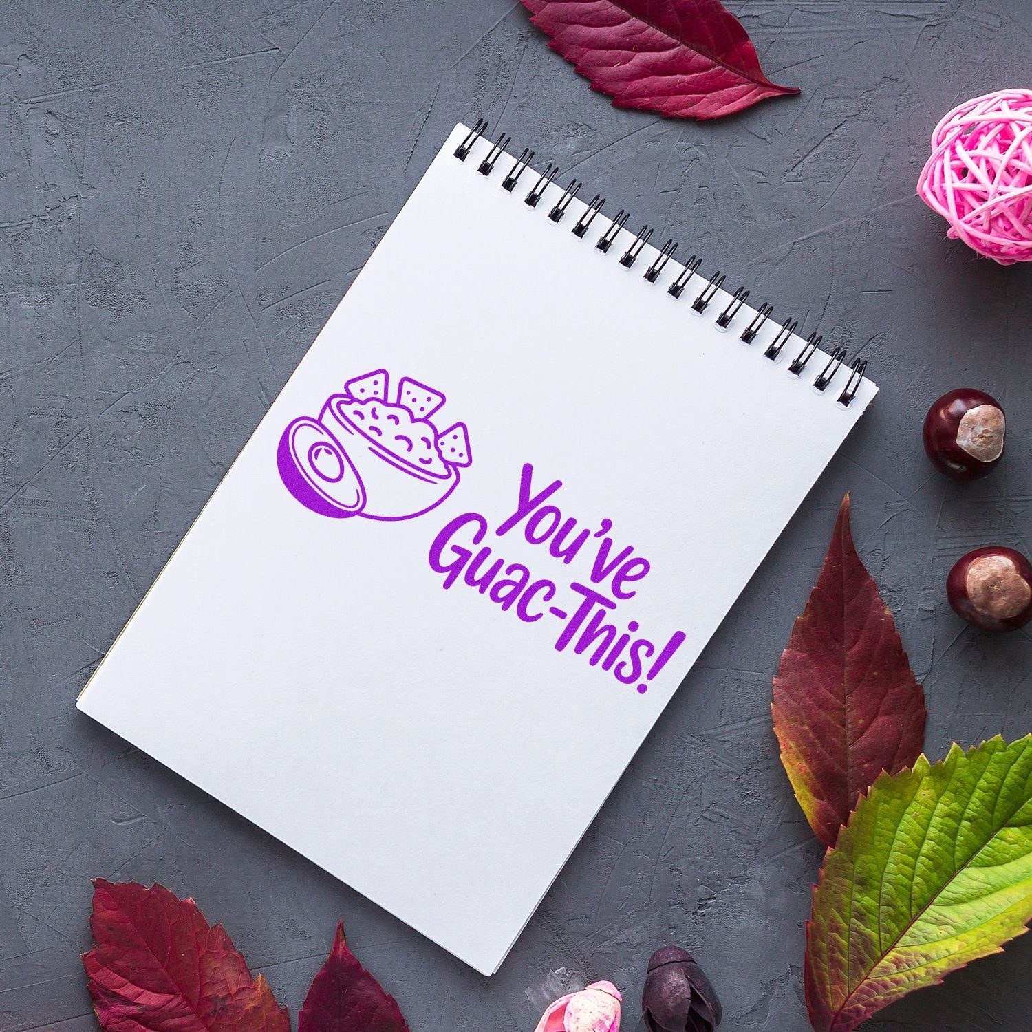 Purple guacamole stamp seal on a white spiral notebook, with avocado bowl icon, on a gray background with autumn leaves.