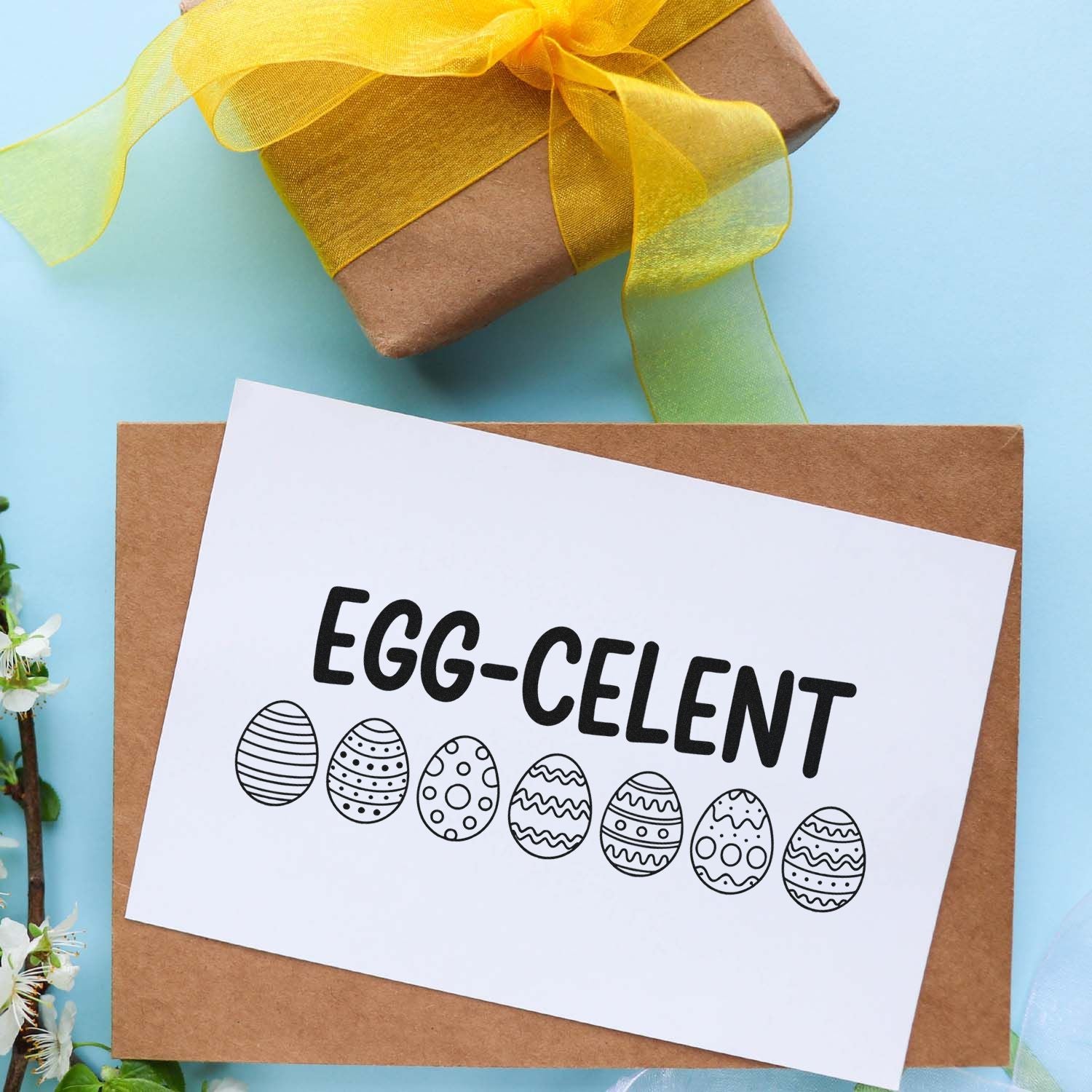 Brown kraft gift box with yellow ribbon and white custom stamp card reading EGG-CELENT with patterned Easter eggs.