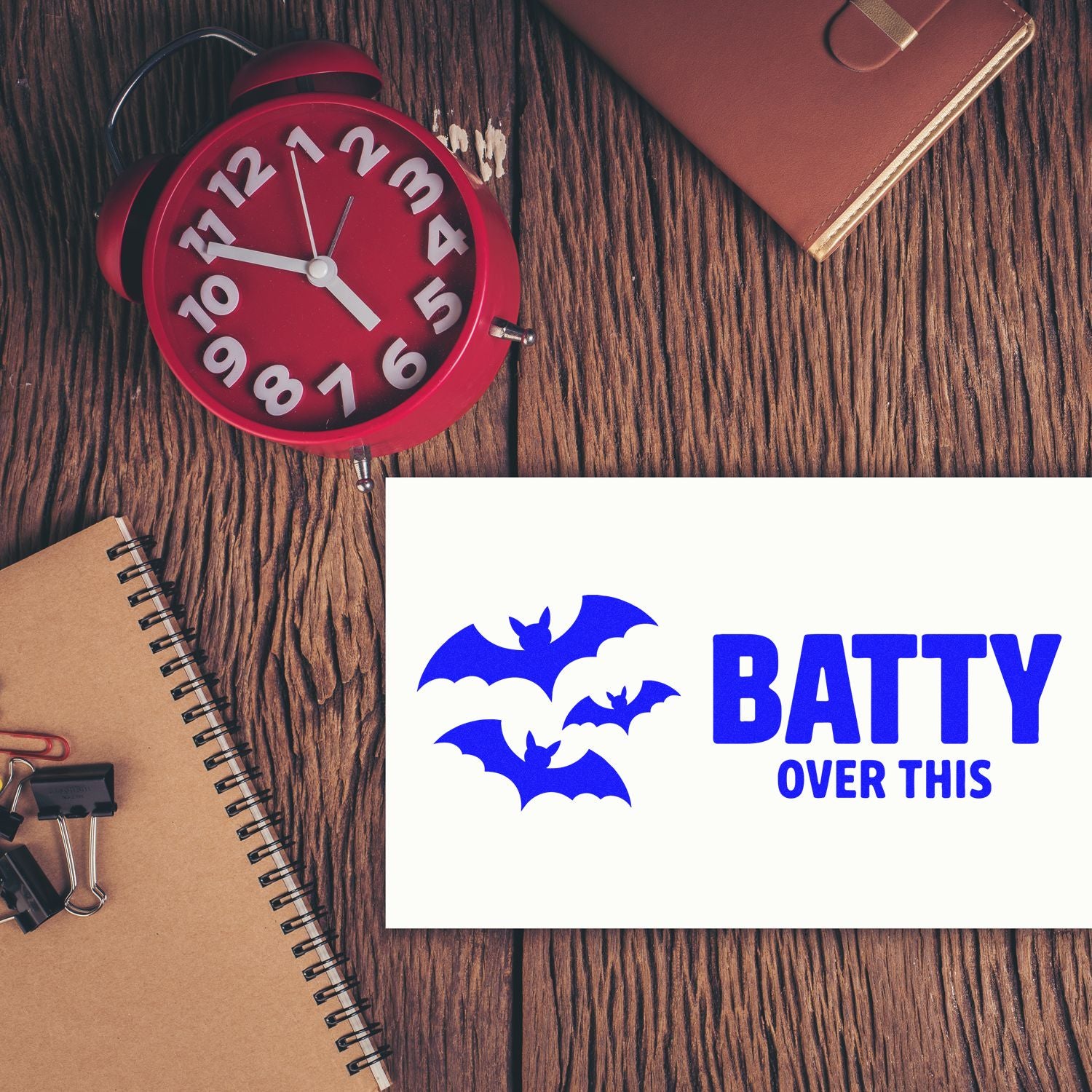 Blue bat-themed stamp seal reading BATTY OVER THIS on a white card beside a red alarm clock.