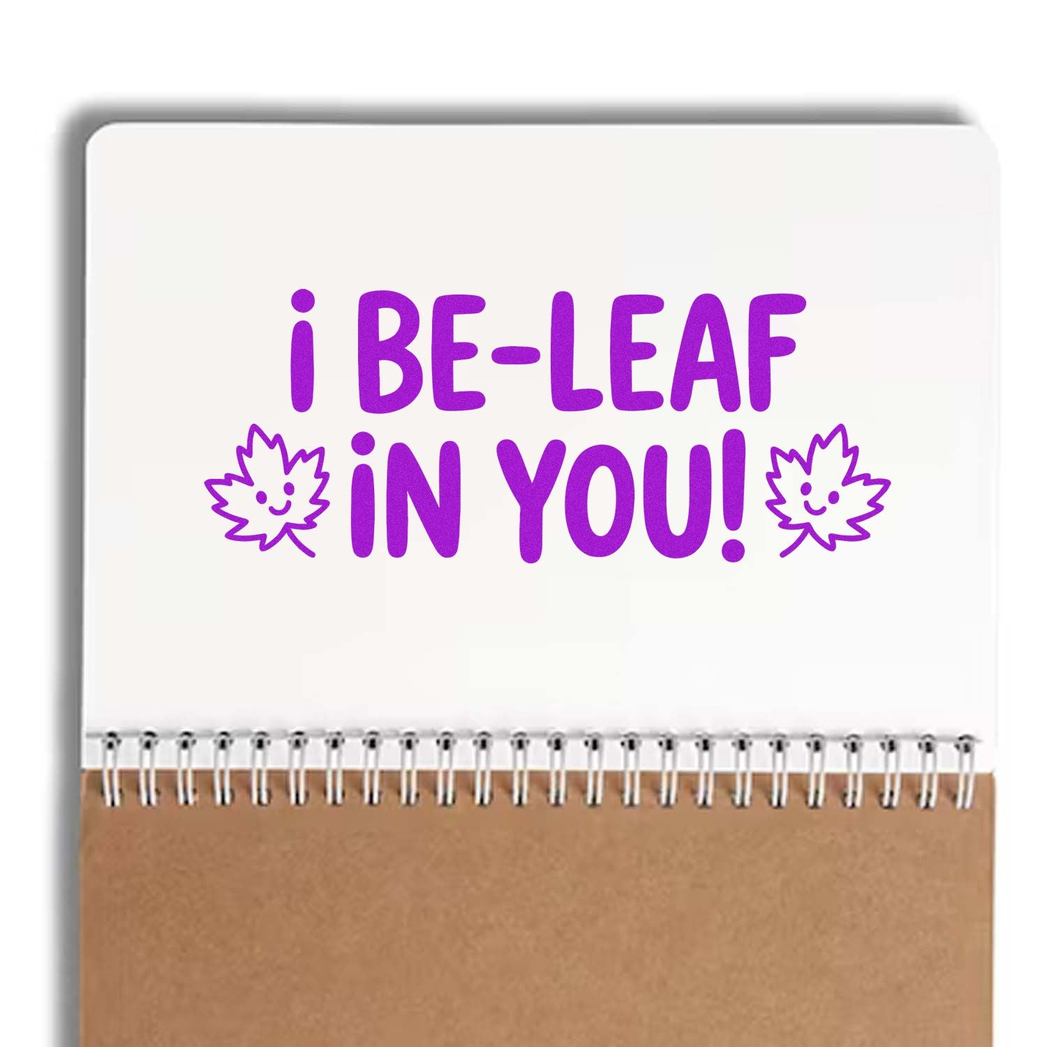 White stamp and seal pad with purple I BE-LEAF IN YOU! text, smiling leaf icons, and brown spiral-bound base.