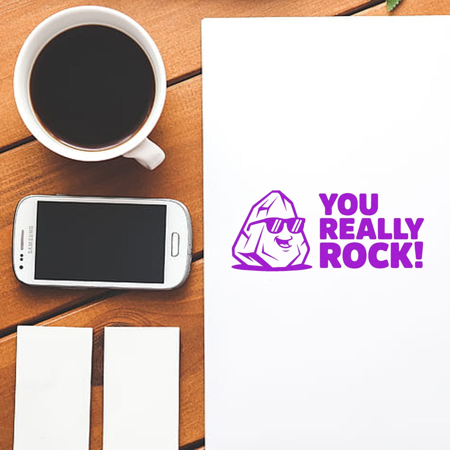 Purple rock mascot stamp and seal on white sheet, reading YOU REALLY ROCK!, beside a coffee cup and smartphone on wood.