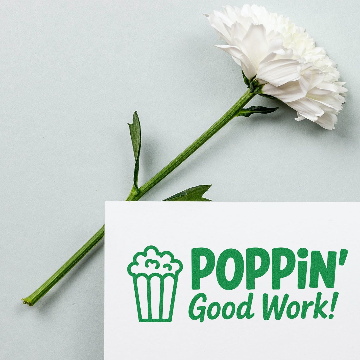 Green stamp and seal reading POPPIN Good Work! with popcorn icon, beside a white flower on a pale gray background.