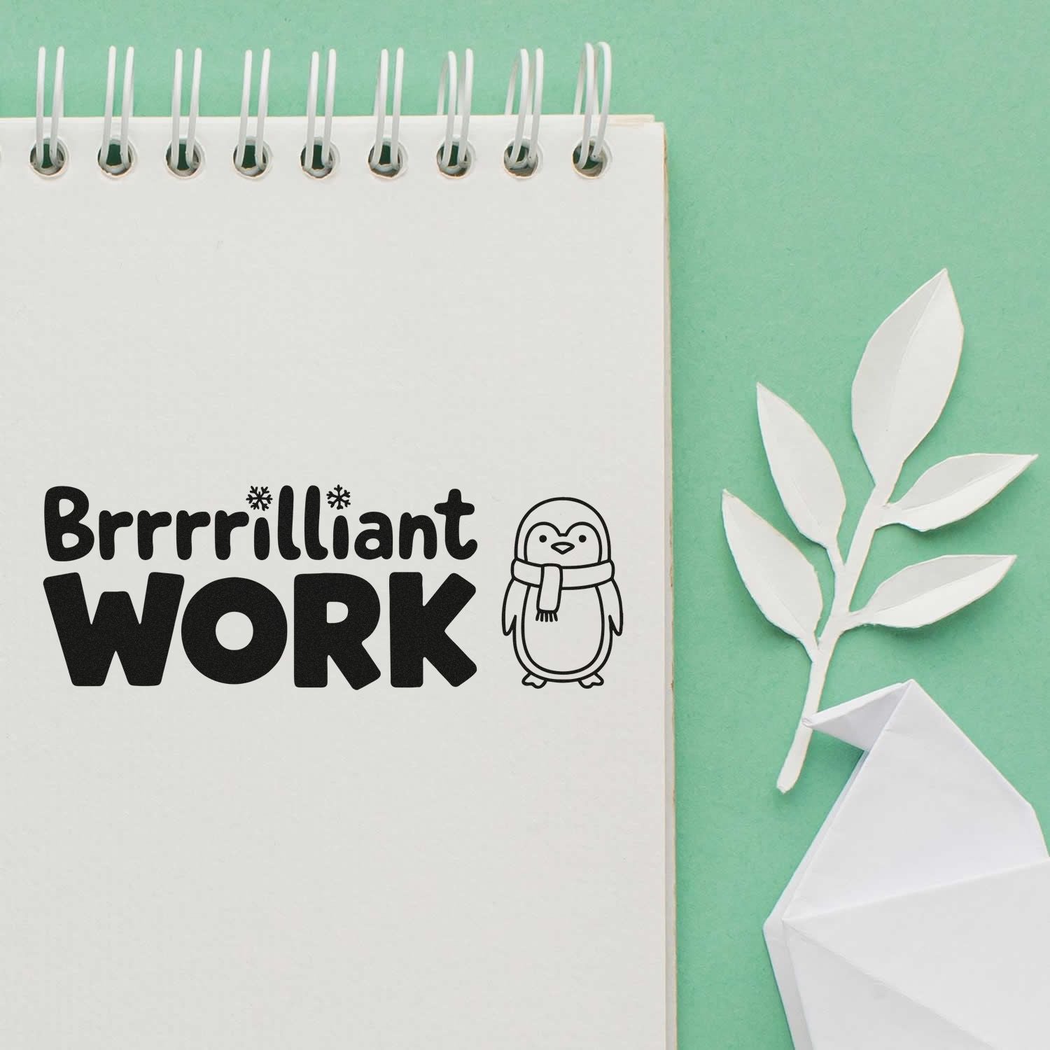 Black Brrrilliant Work stamp with penguin seal on white spiral notebook, on mint green background with white paper leaf.
