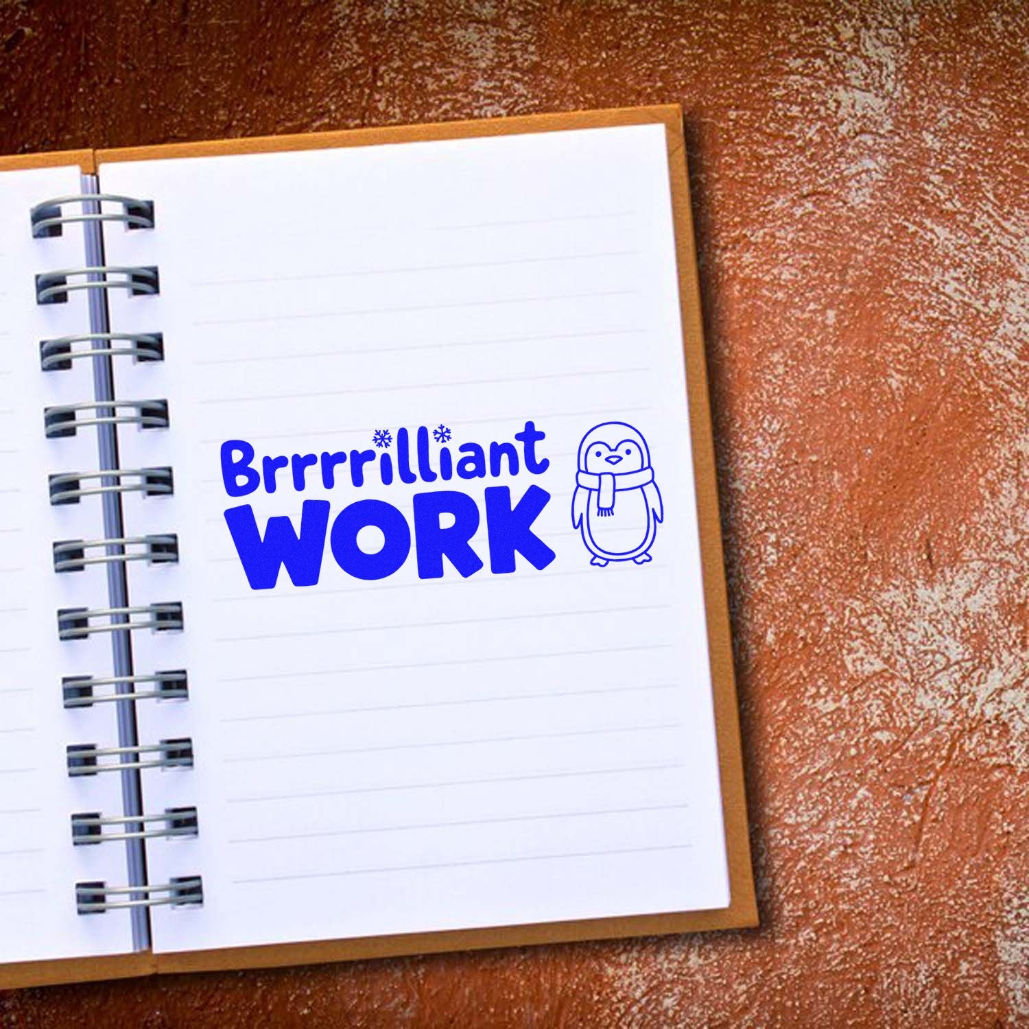 Blue Brrrrilliant Work penguin stamp seal on a white spiral notebook, set on a brown textured background.
