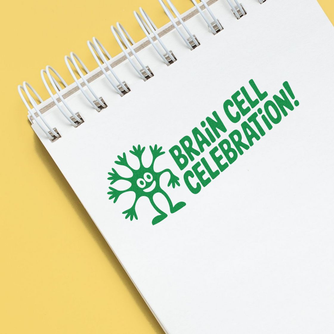 Green Brain Cell Celebration stamp with cartoon neuron on a white spiral notebook over a yellow background.