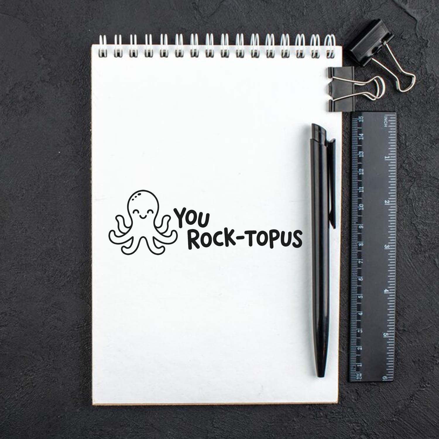 Black octopus custom stamp reading You Rock-topus on white spiral notebook with black pen, ruler, and binder clips.