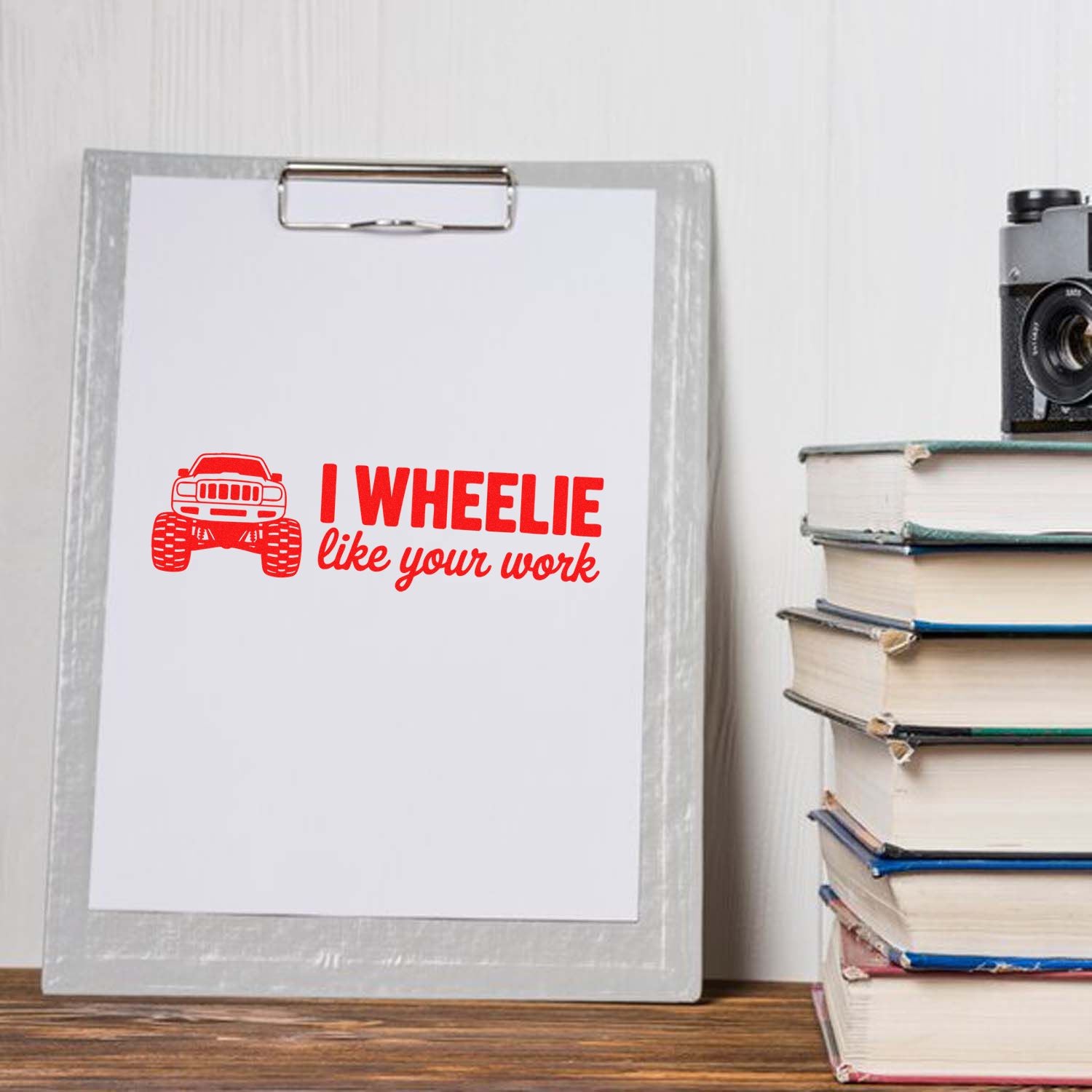 Red monster truck stamp and seal design with I wheelie like your work on clipboard beside stacked books.