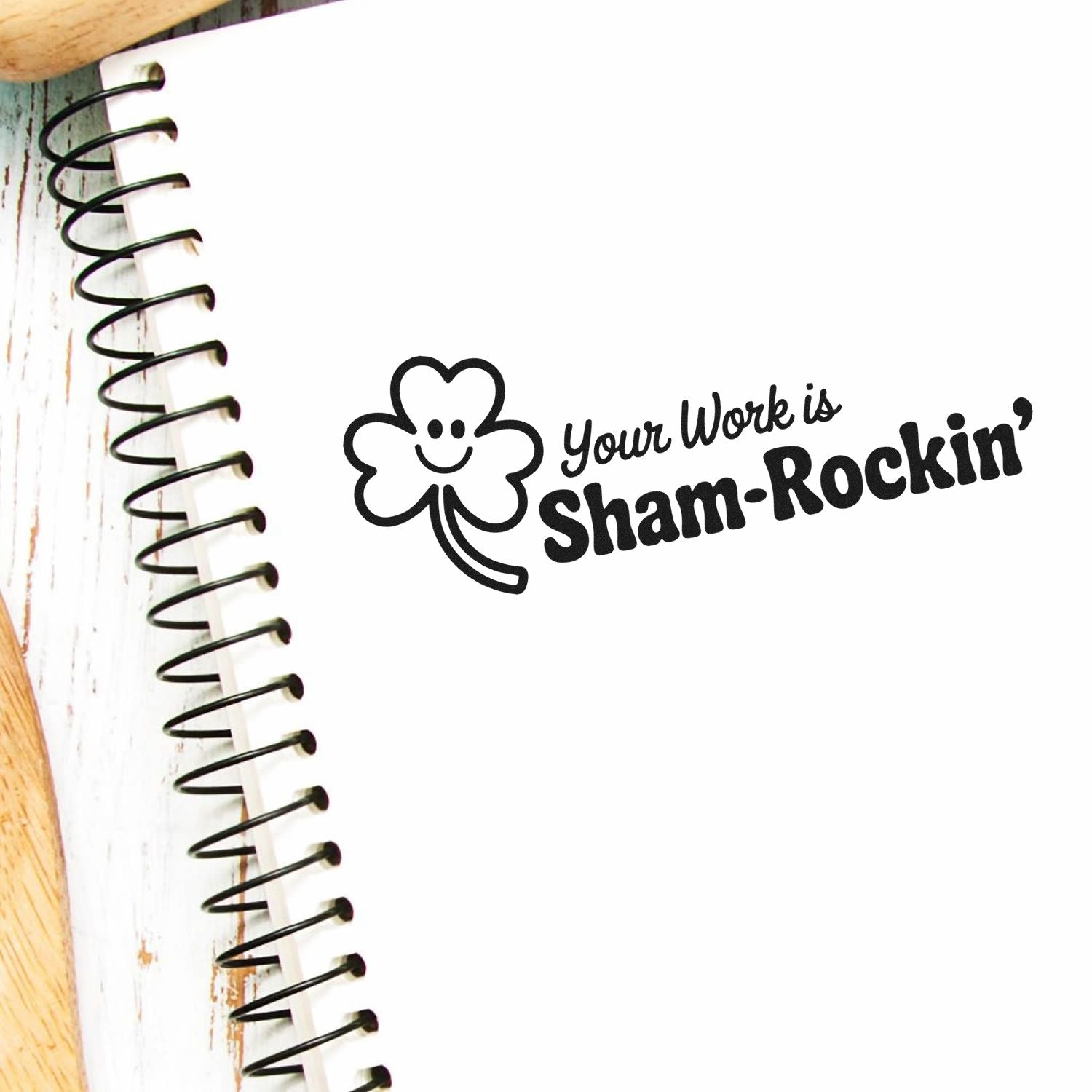 Black Sham-Rockin stamp seal with smiling clover on white notebook, beside spiral binding and wood edge.