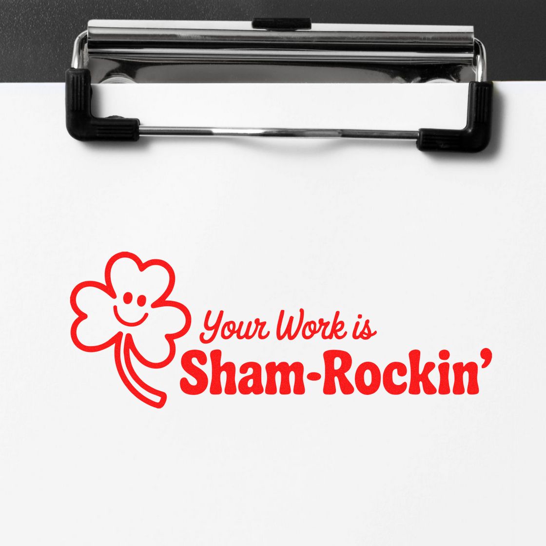 Red shamrock rubber stamp design on white paper clipped to a black clipboard, reading Your Work is Sham-Rockin.