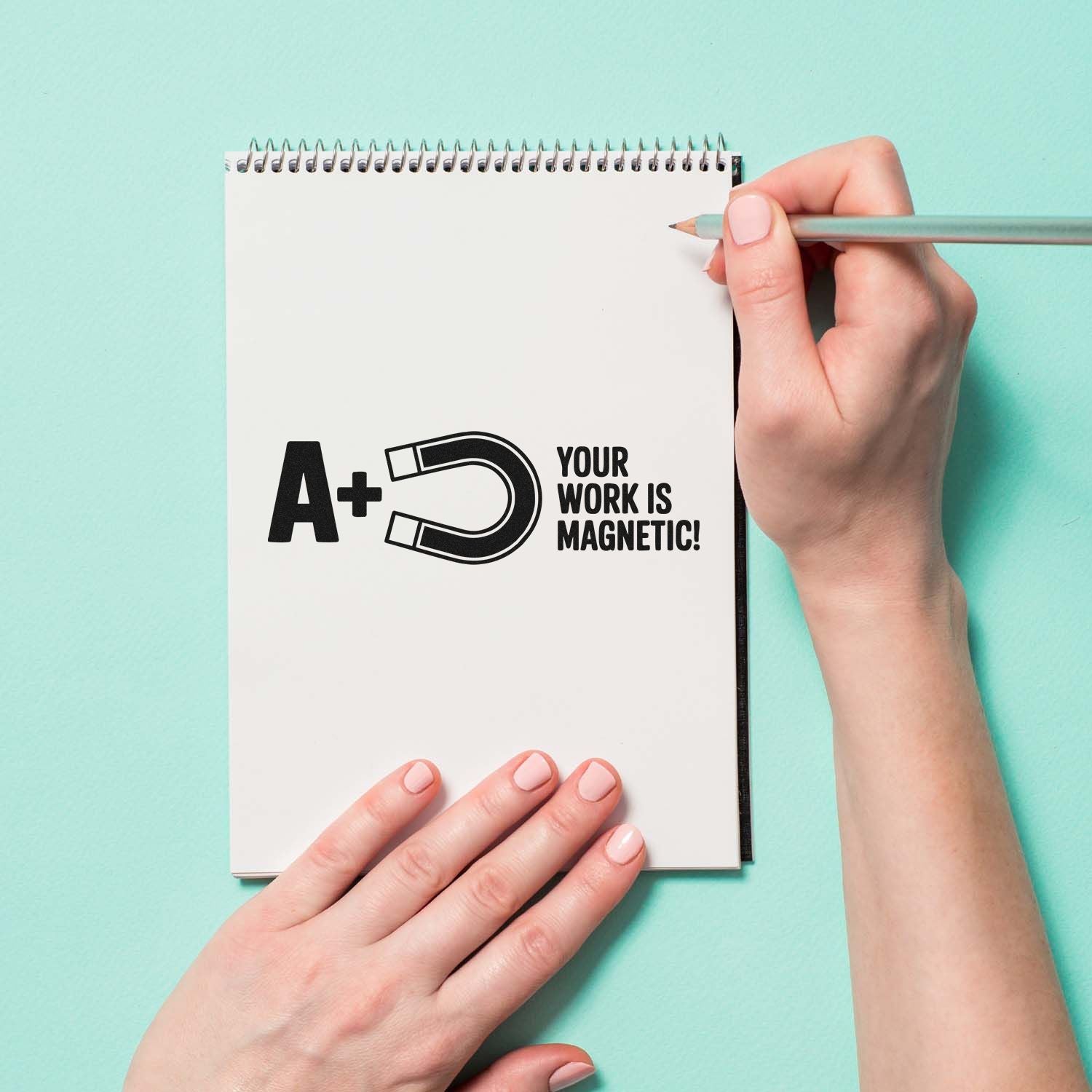 Mint-green background with notebook featuring A+ magnet stamp seal, Your work is magnetic! text, and a hand holding a pencil.