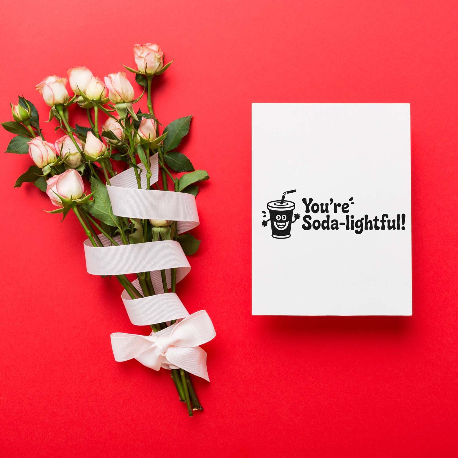 Pink roses tied with white ribbon beside a white card stamped Youre Soda-lightful! on a bright red background.