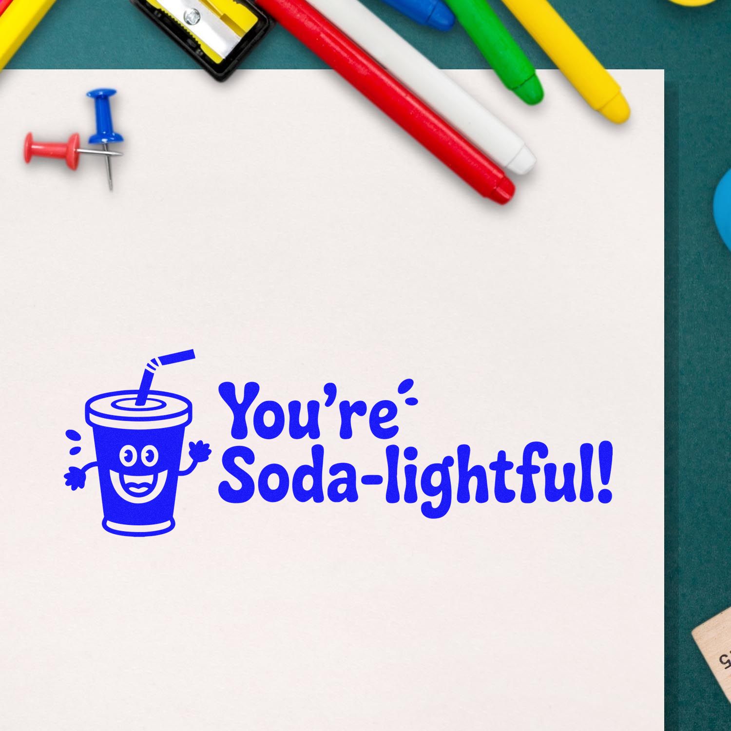 Blue rubber stamp seal with smiling soda cup and Youre Soda-lightful! on white paper, with colorful pens and push pins.