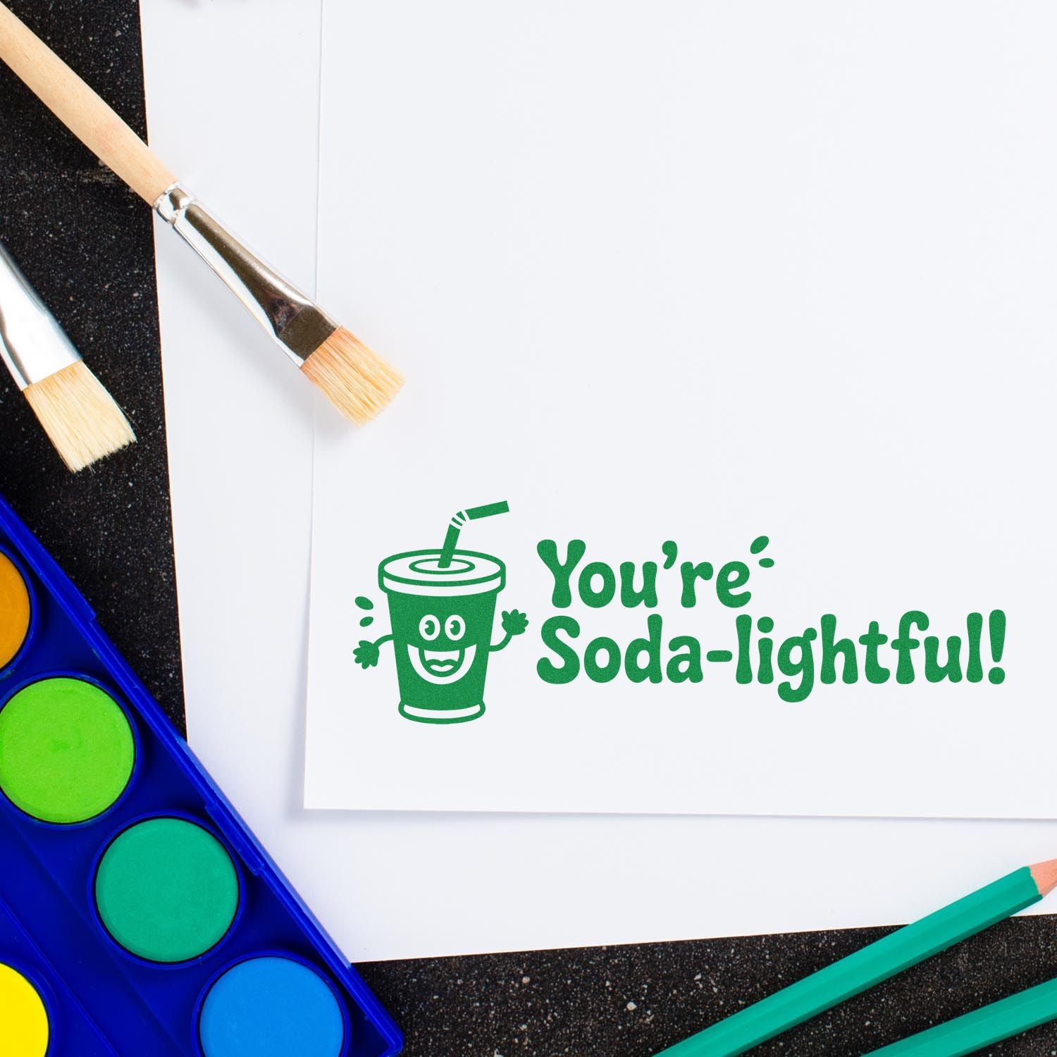 Green soda cup stamp and seal on white paper with Youre Soda-lightful! beside paintbrushes and watercolor paints.