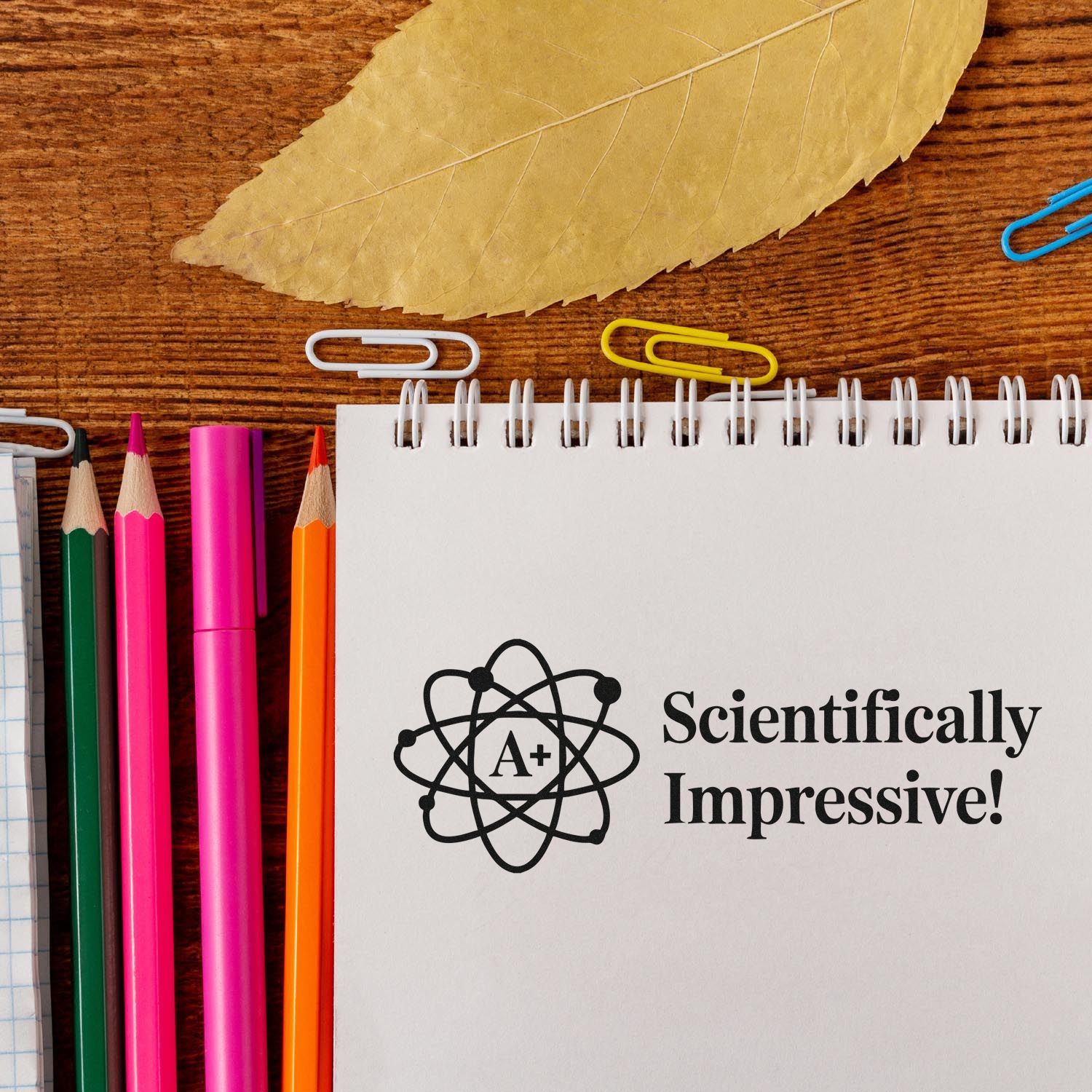 Scientifically Impressive! A+ atom stamp seal on spiral notebook, with pink, green, orange pencils and colorful paper clips.