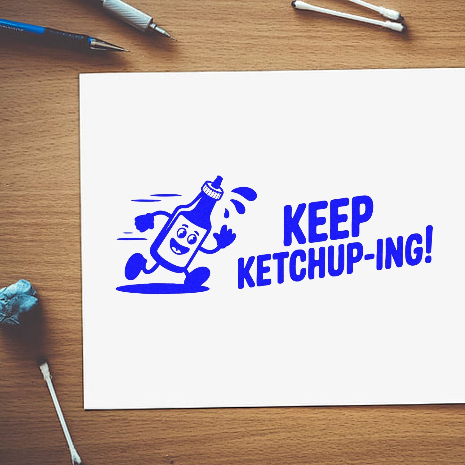 Blue rubber stamp design of a smiling ketchup bottle with KEEP KETCHUP-ING! on white paper.
