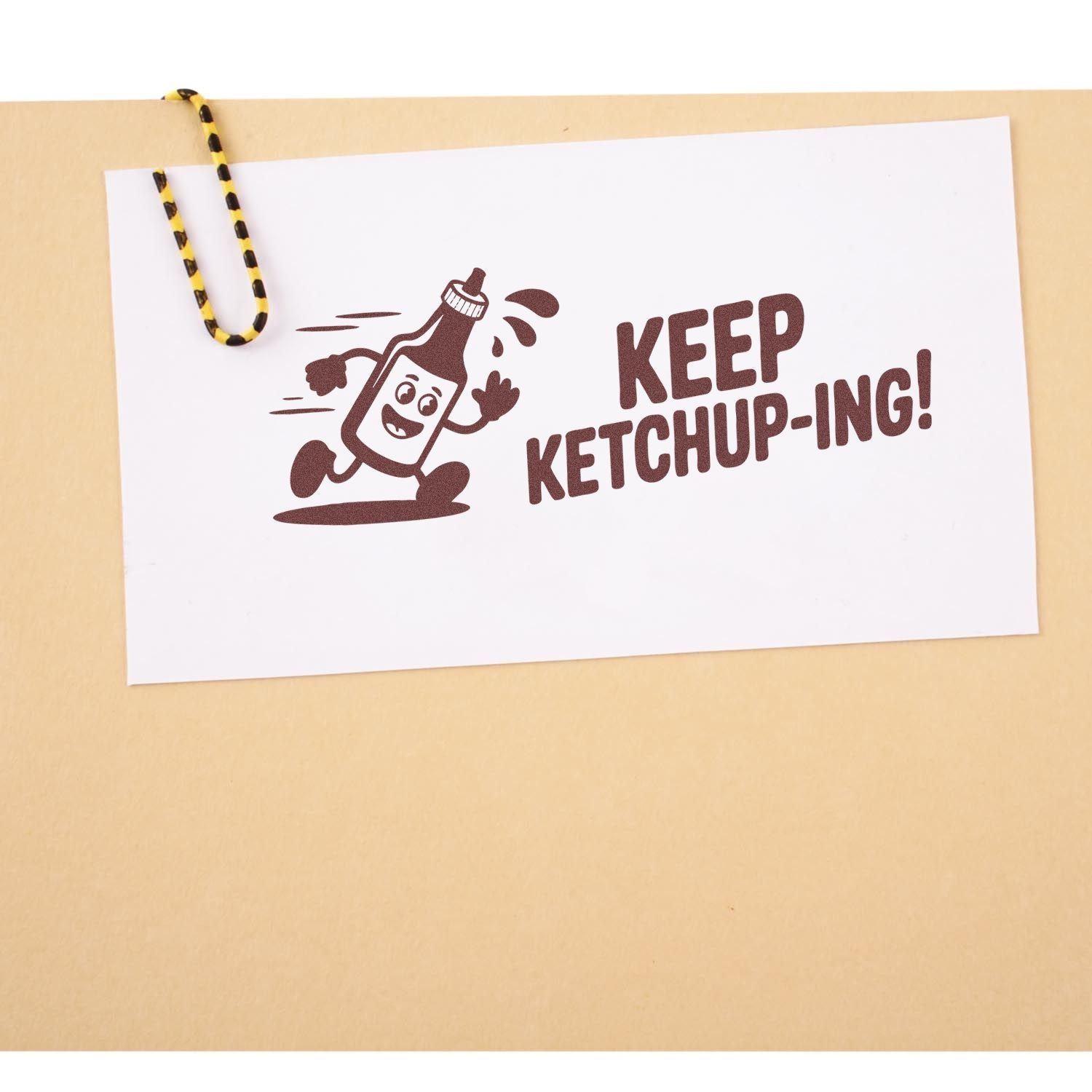 Brown ketchup bottle stamp and seal reading KEEP KETCHUP-ING! on white card, clipped with a yellow-black paperclip.