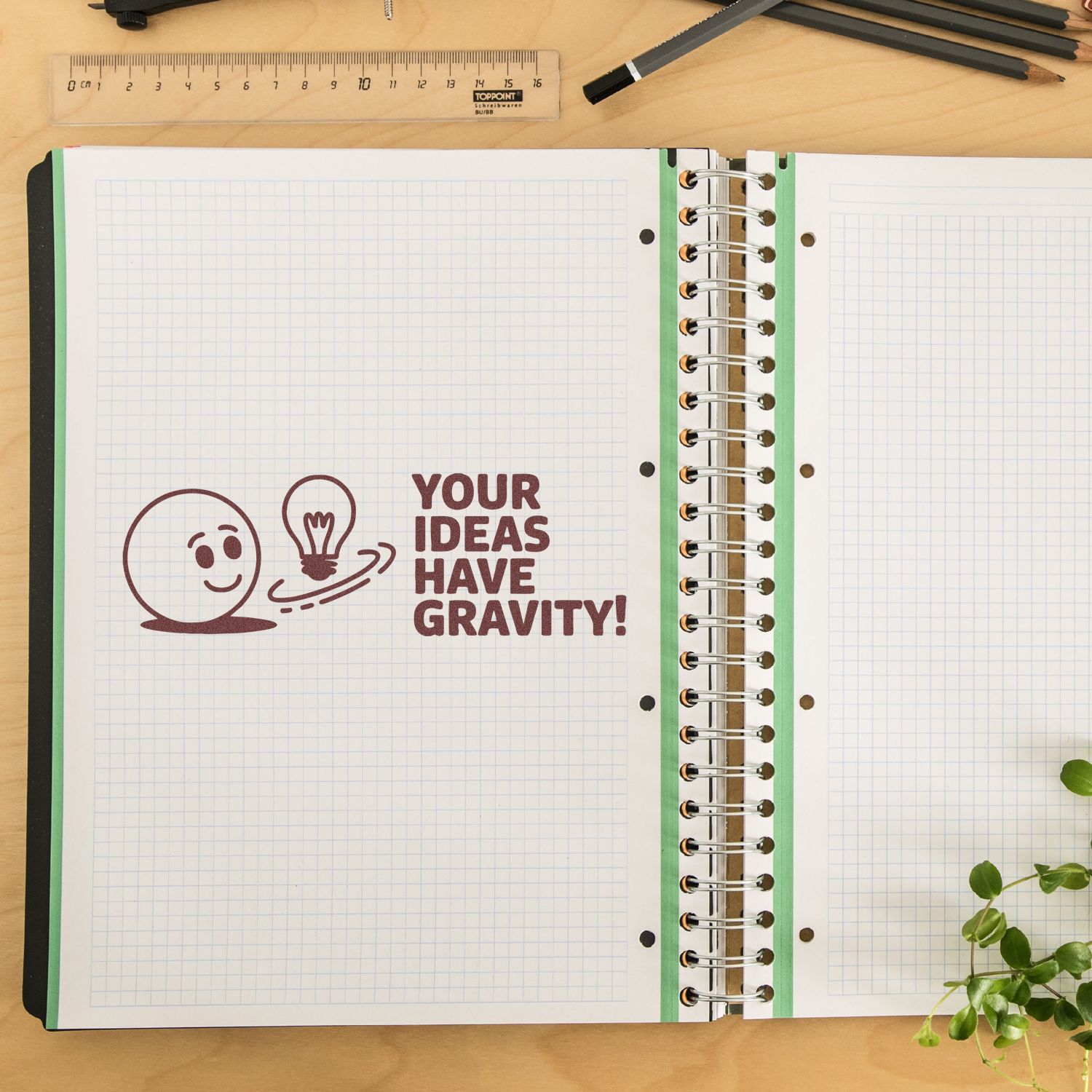 Maroon rubber stamp on open grid notebook says Your ideas have gravity! with smiley face, lightbulb, and spiral rings.