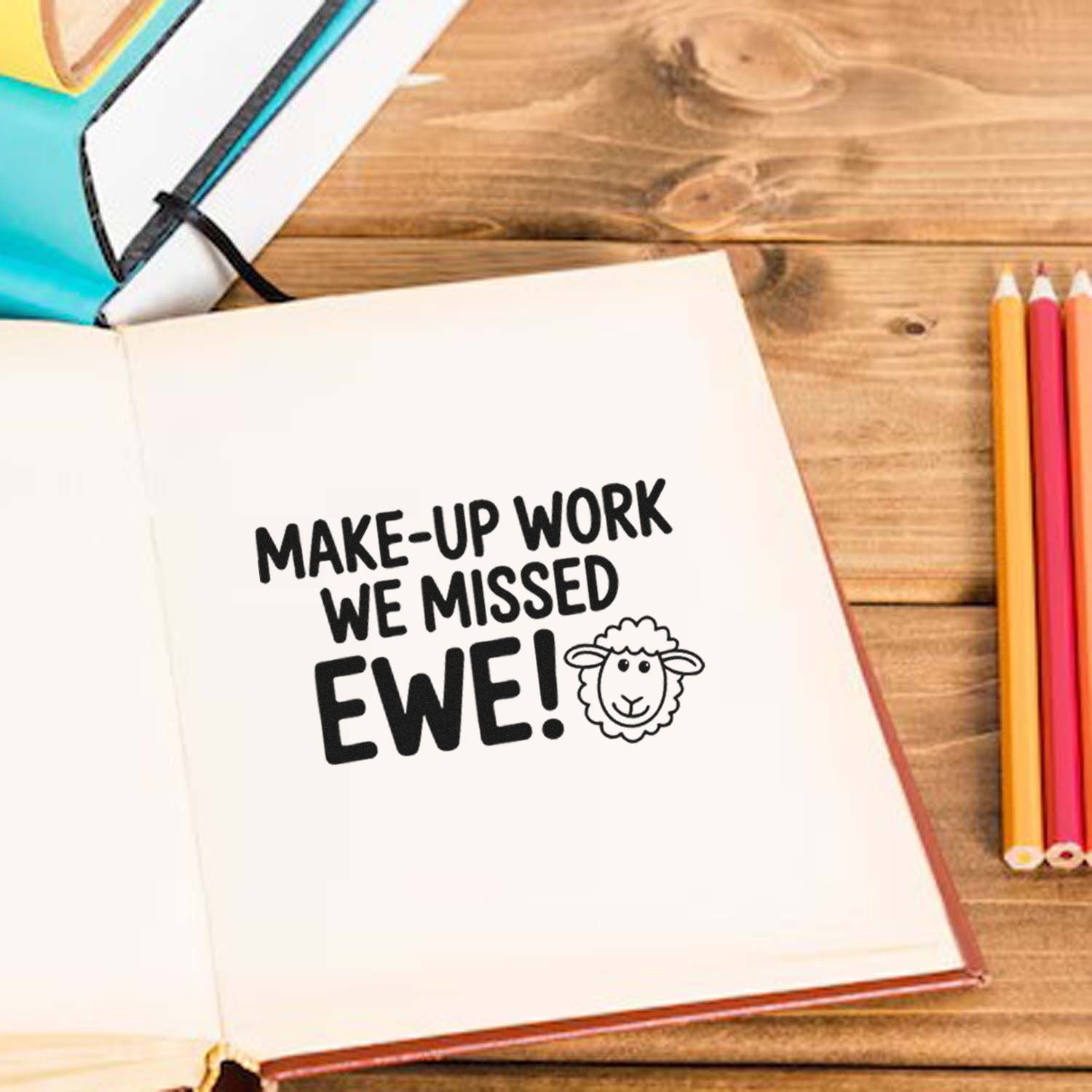 Black custom rubber stamp on white notebook reads MAKE-UP WORK WE MISSED EWE! beside colorful pencils on a wood desk.
