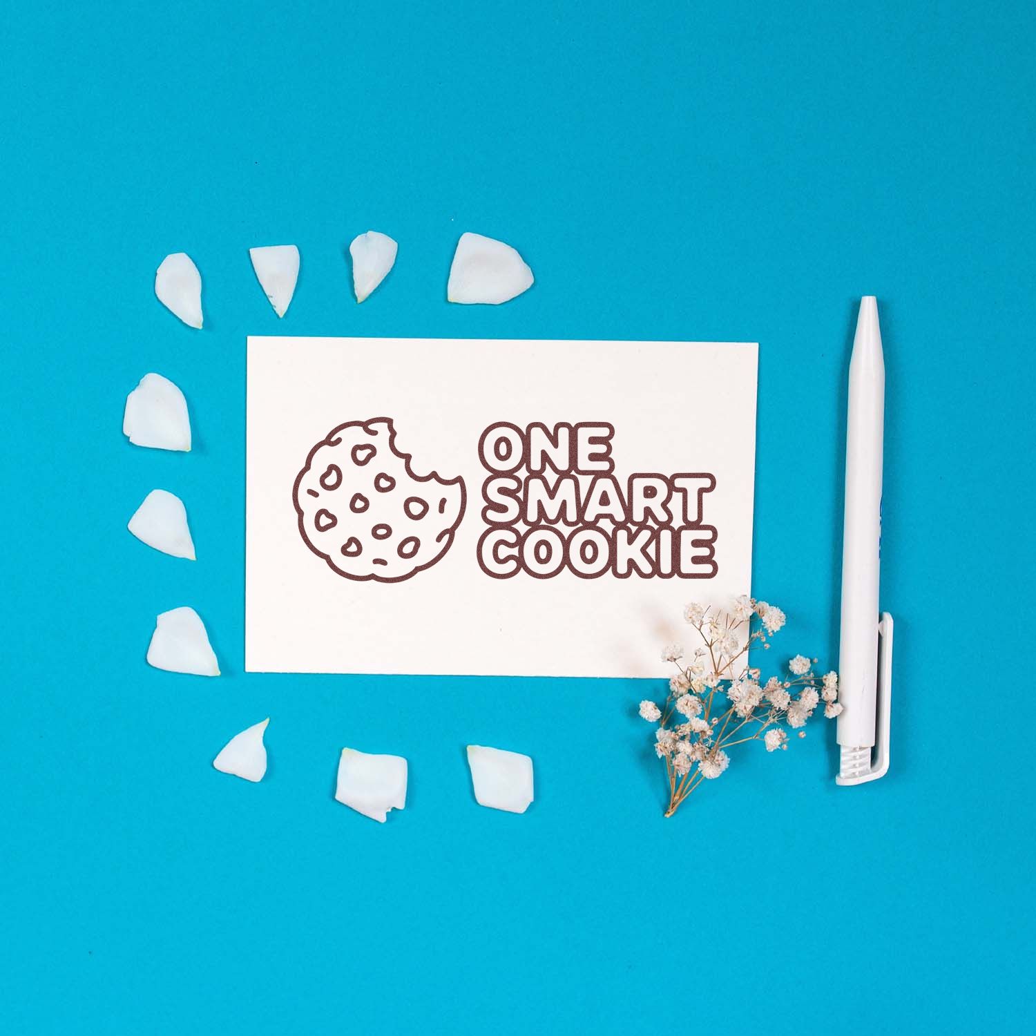 White cookie stamp and seal card reading ONE SMART COOKIE on bright blue background, with white petals and a white tool