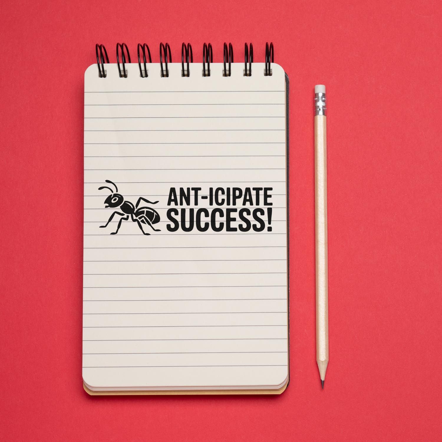 Black rubber ant success stamp on a lined notebook beside a pencil, on a bright red background.