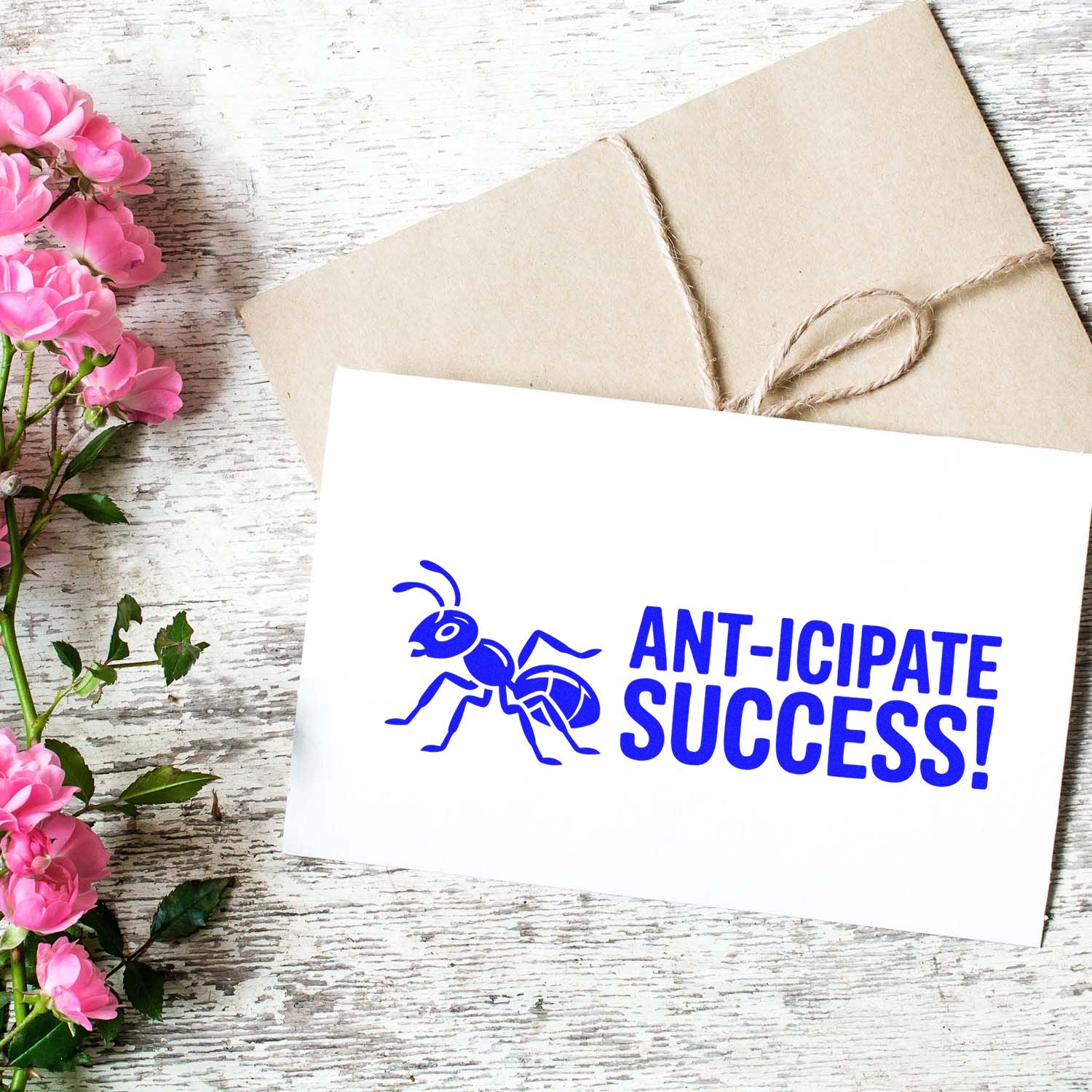 Blue custom stamp and seal with ant graphic and ANT-ICIPATE SUCCESS! on white card, beside kraft envelope and pink flowers.