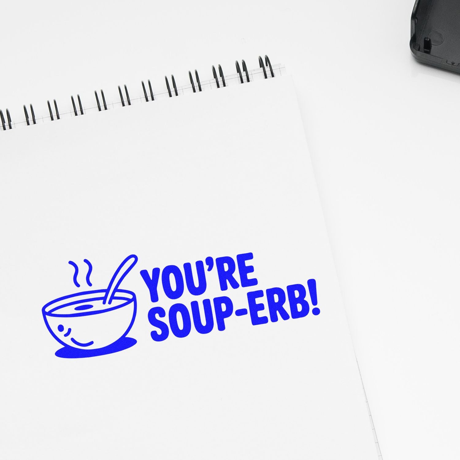 Blue rubber stamp reading YOURE SOUP-ERB! with a smiling soup bowl icon on a white spiral notebook.