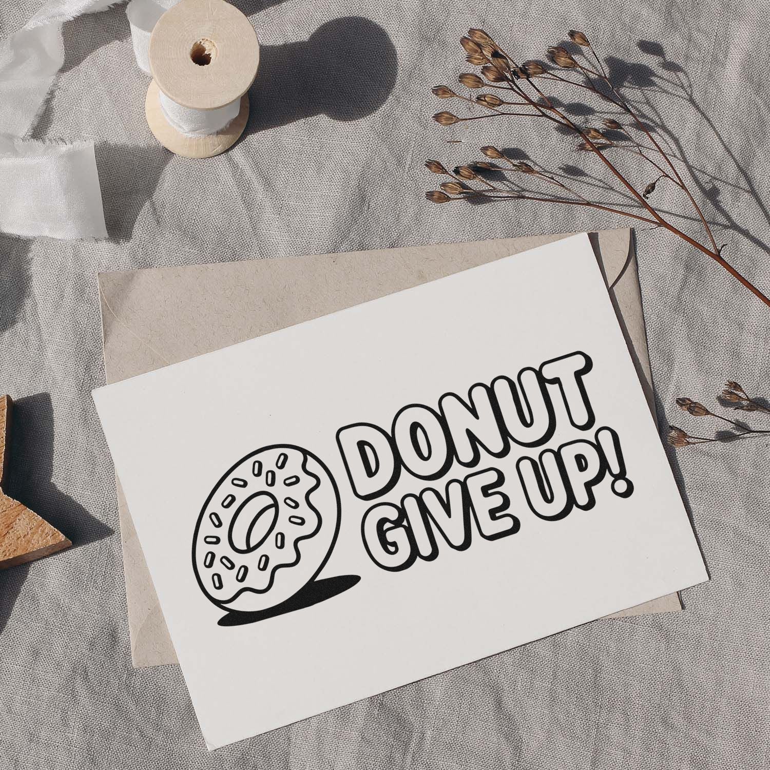 Black donut-shaped stamp and seal reading DONUT GIVE UP! on a white card, with beige envelope, ribbon spool, and dried stems.