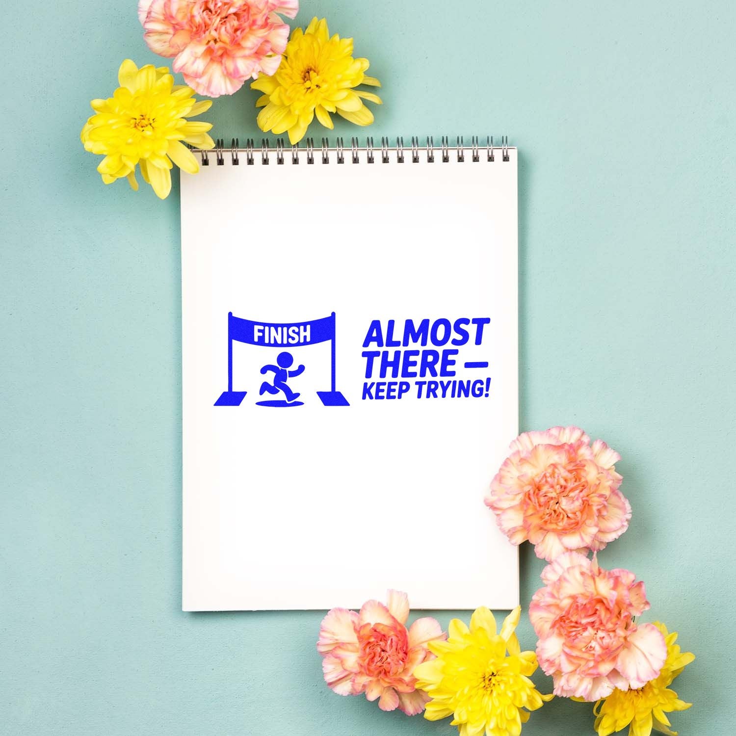 Blue finish-line motivational stamp and seal on white spiral notebook, surrounded by pink and yellow flowers on mint background.
