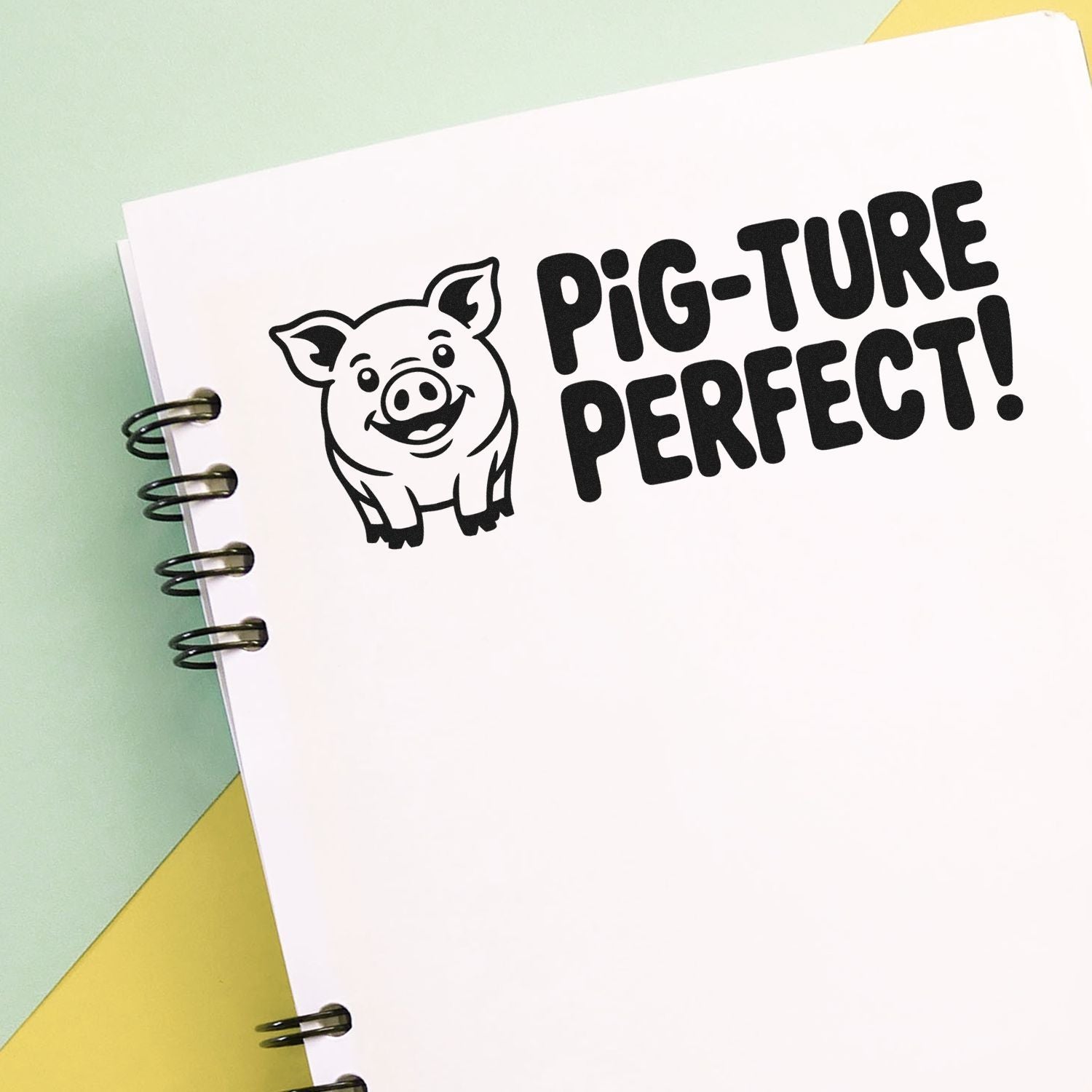 Black pig stamp seal reading PIG-TURE PERFECT! on white spiral notebook, with cute pig icon and pastel green-yellow background.