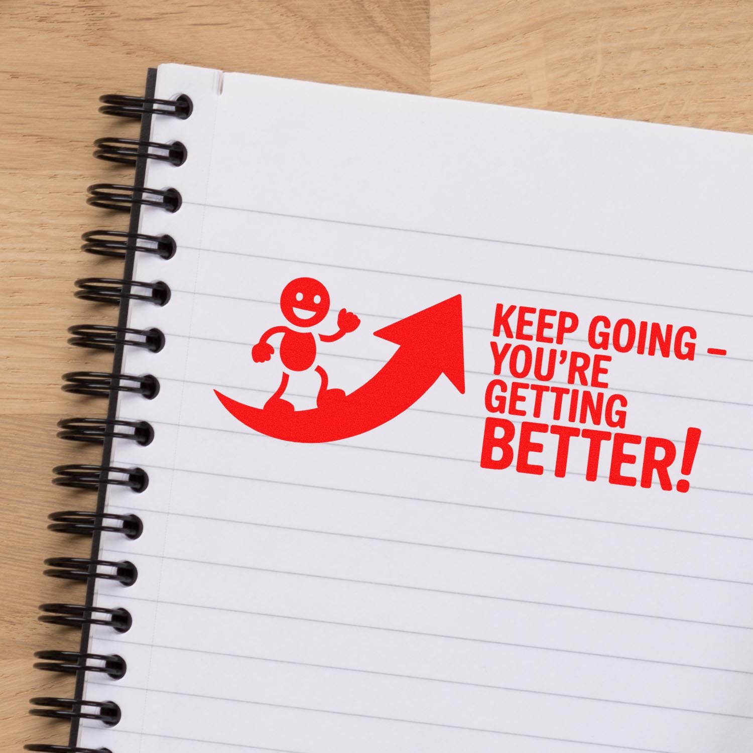 Red self-inking stamp seal on a lined spiral notebook, featuring a smiling figure, upward arrow, and Keep goingyoure getting better!