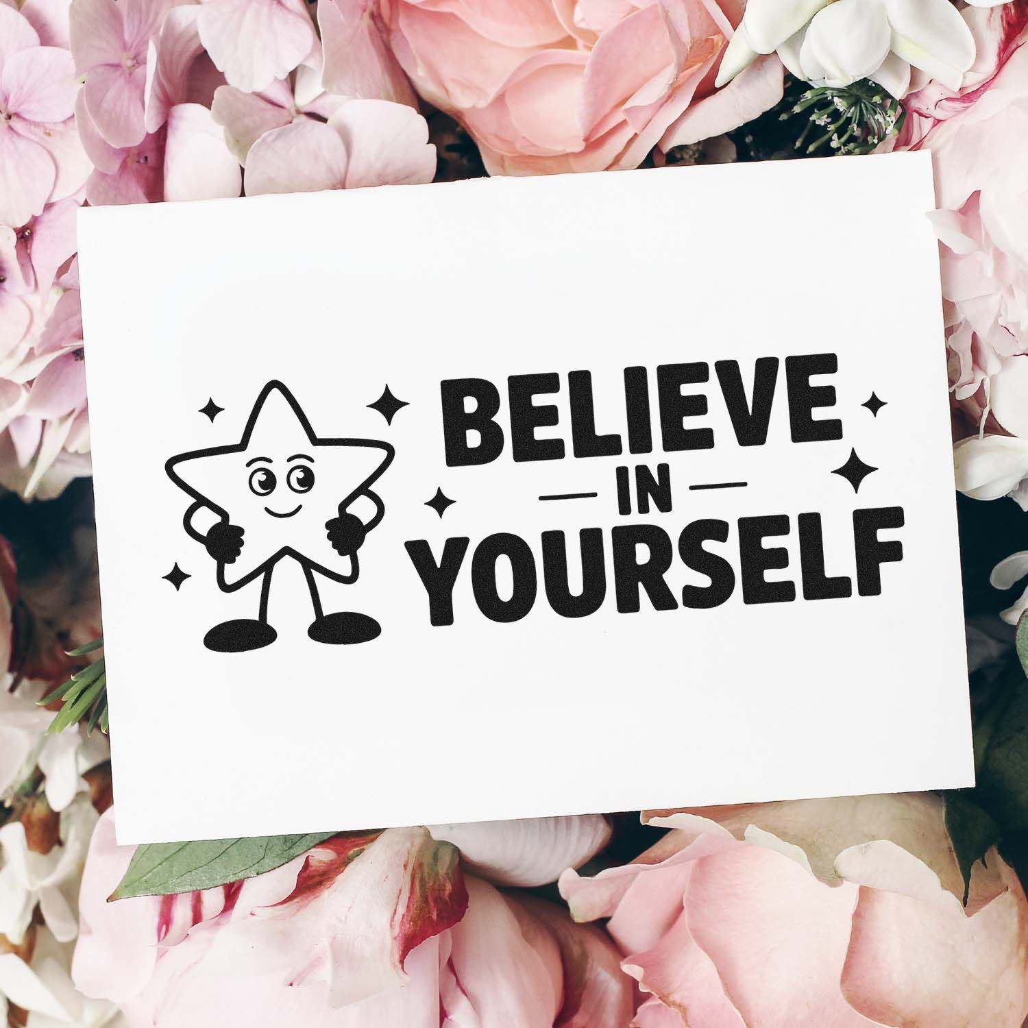 Black stamp and seal design with smiling star and Believe in Yourself on a white card, surrounded by soft pink flowers.