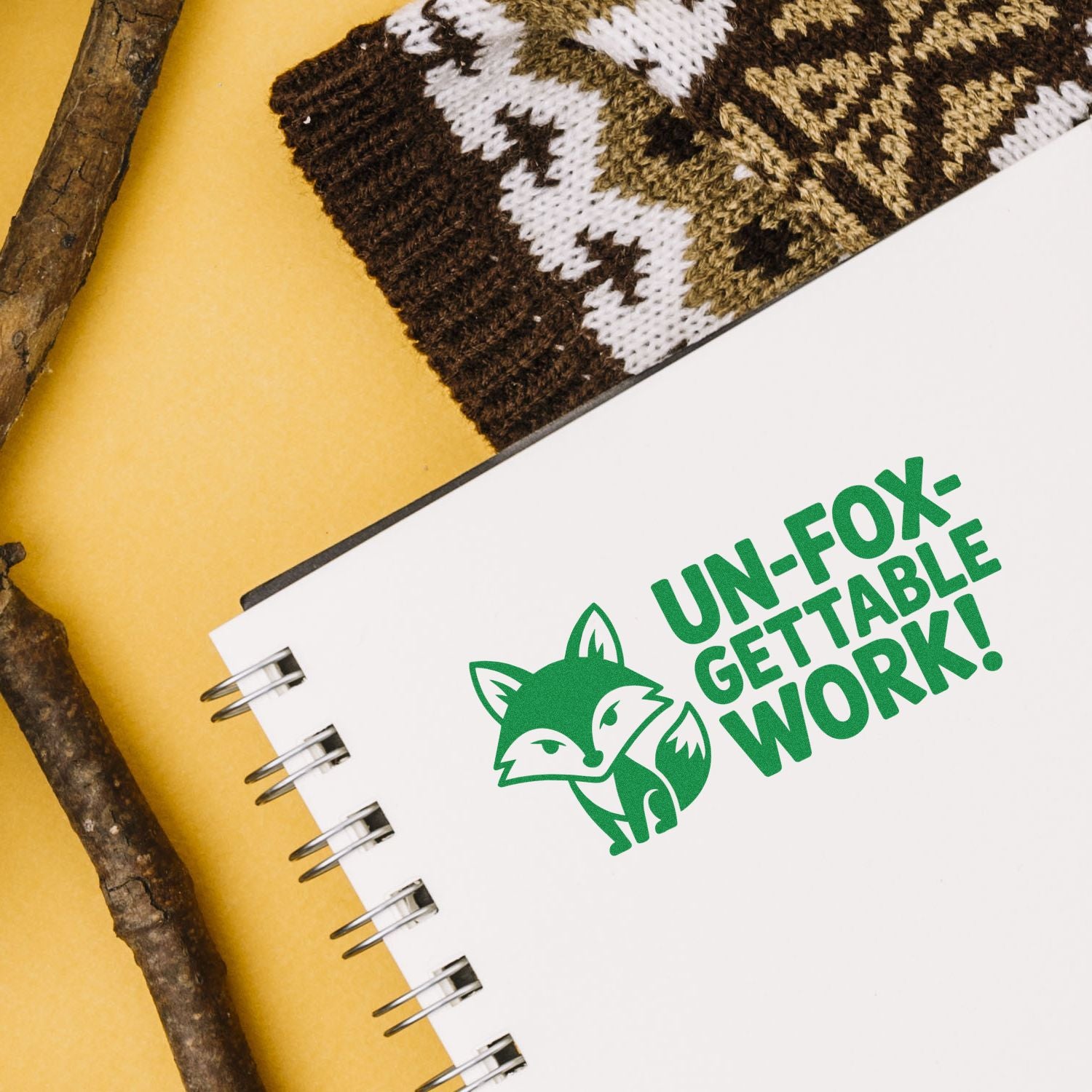 Green fox logo stamp on white spiral notebook, with UN-FOX-GETTABLE WORK! seal on yellow background and brown knit fabric.