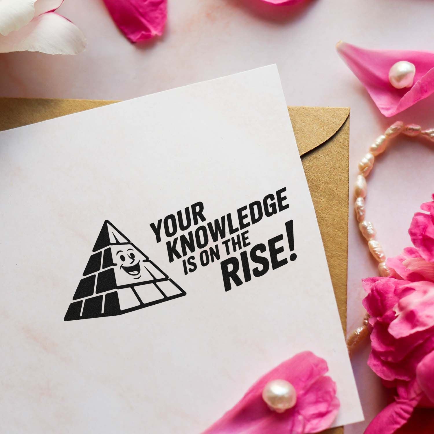 White card with black pyramid stamp reading Your knowledge is on the rise! over a brown envelope, with pink petals and pearls.