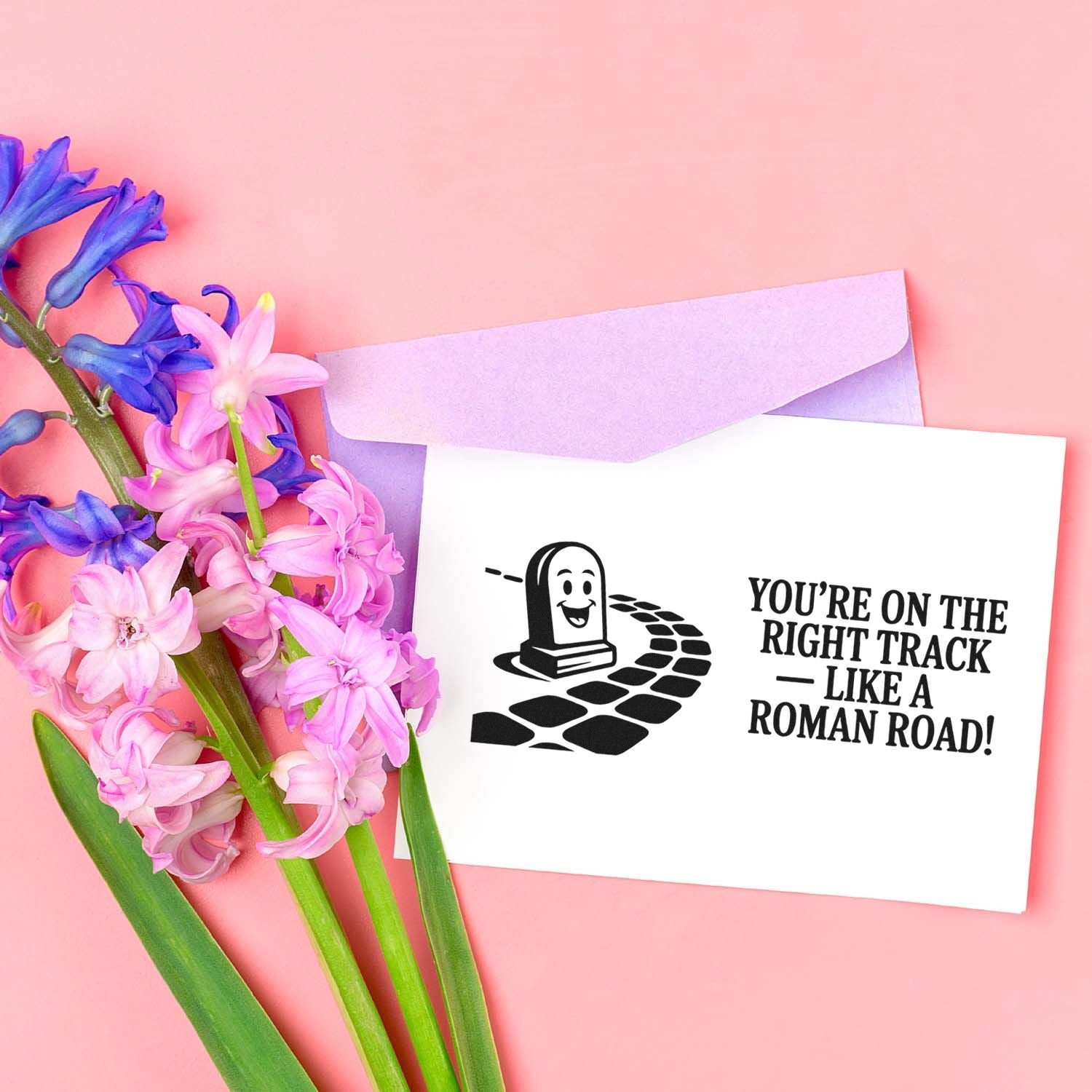 Black stamp seal card with Youre on the right track like a Roman road! beside lilac envelope and pink-purple flowers on pink background.