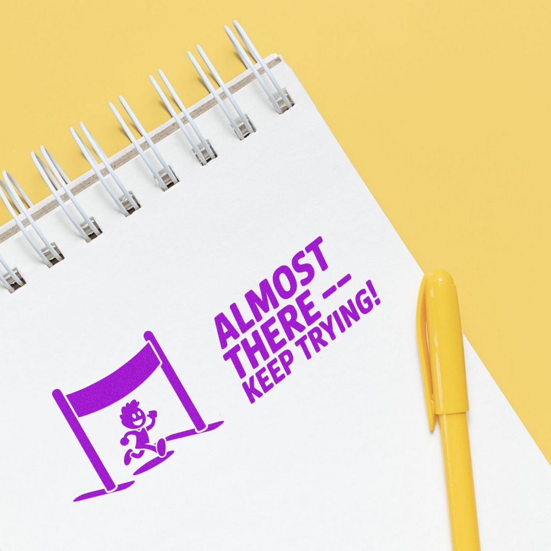 Purple Almost therekeep trying! stamp seal on a white spiral notebook, with a yellow pen on a pale yellow background.