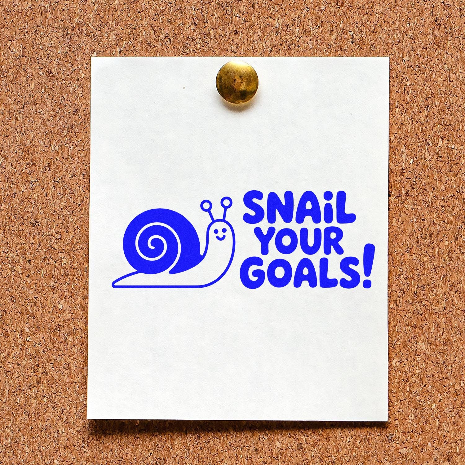 Blue snail stamp and seal design with Snail Your Goals! on white card pinned to corkboard, brass tack.