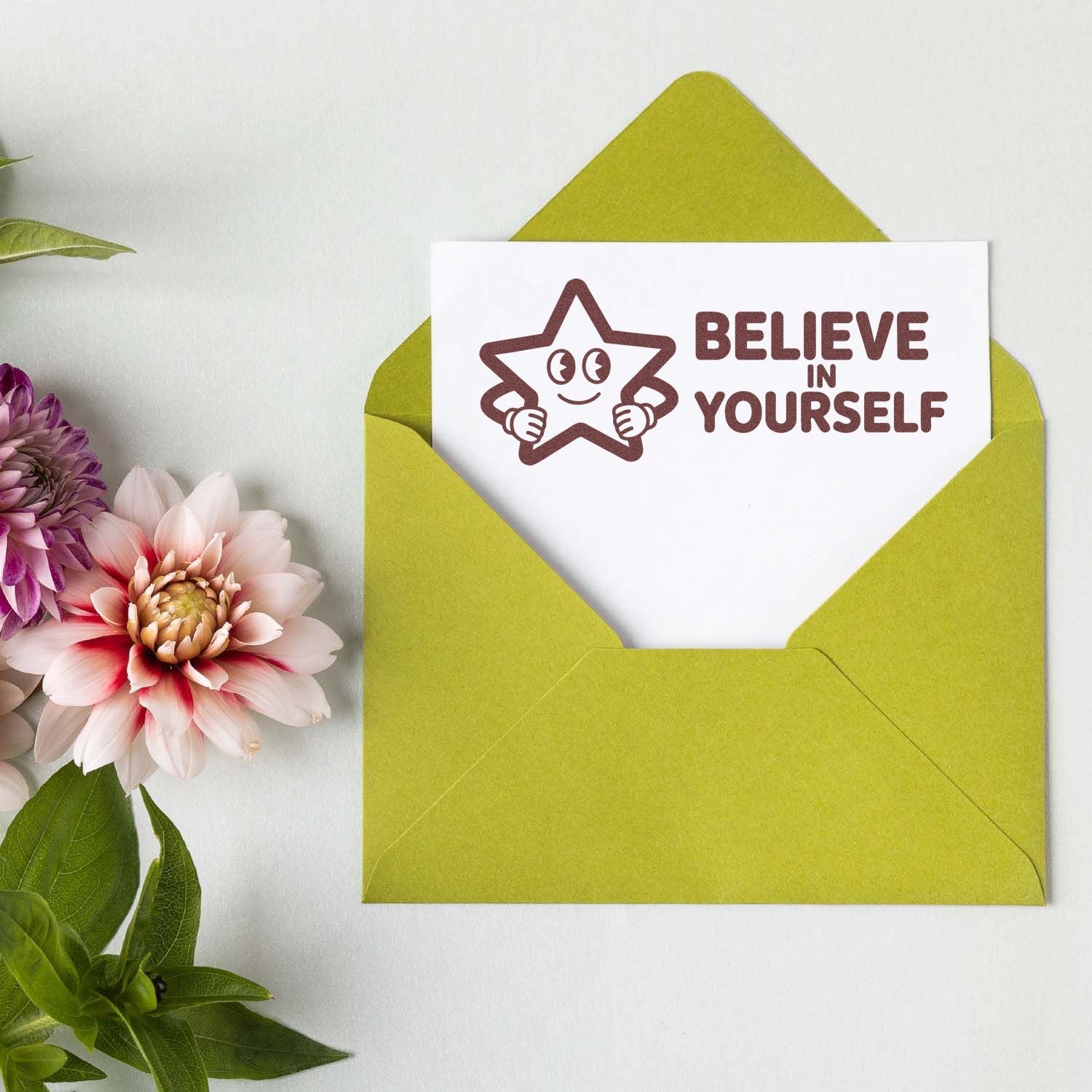 Green envelope with white card featuring brown Believe in Yourself star stamp-and-seal design beside pink flowers.