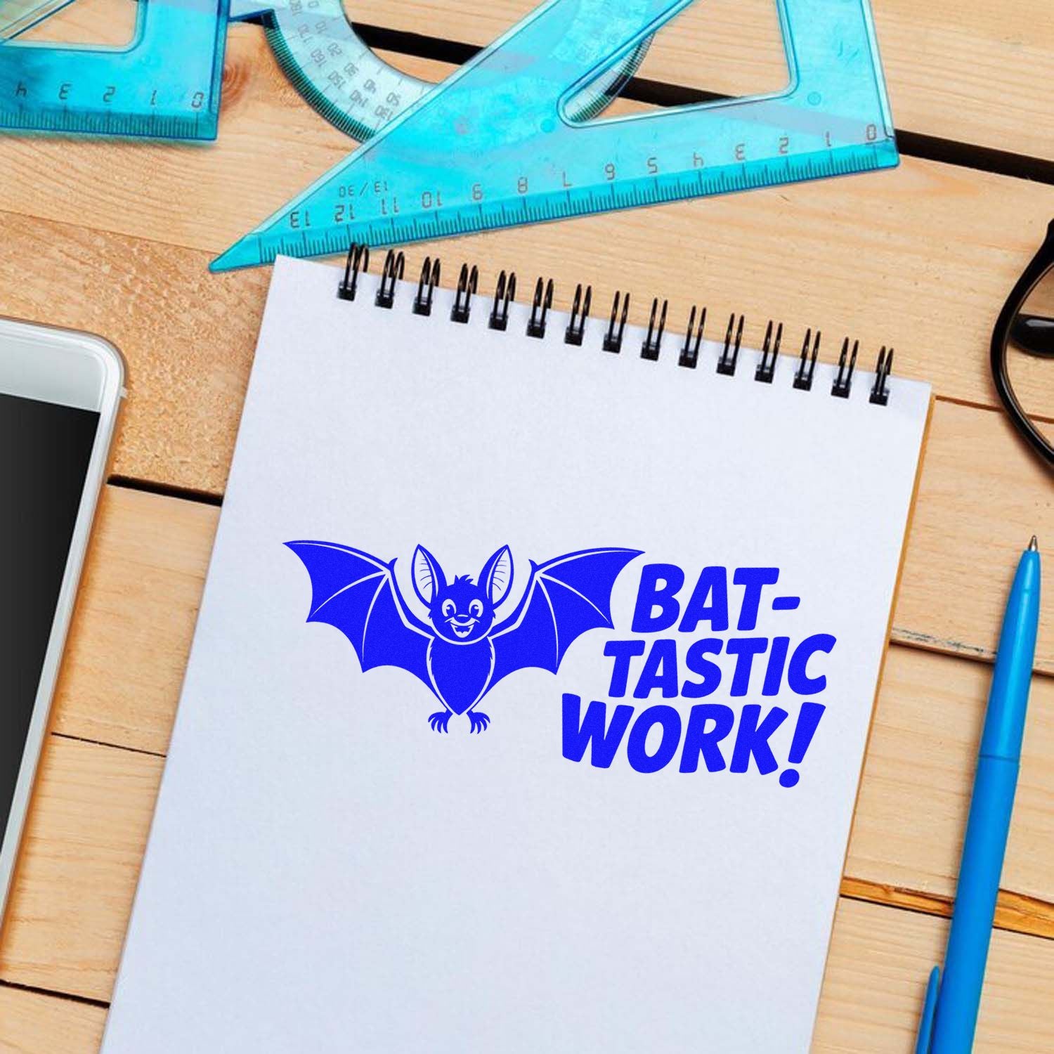 Blue bat stamp and seal with BAT-TASTIC WORK! on a white notebook, surrounded by teal drafting tools.