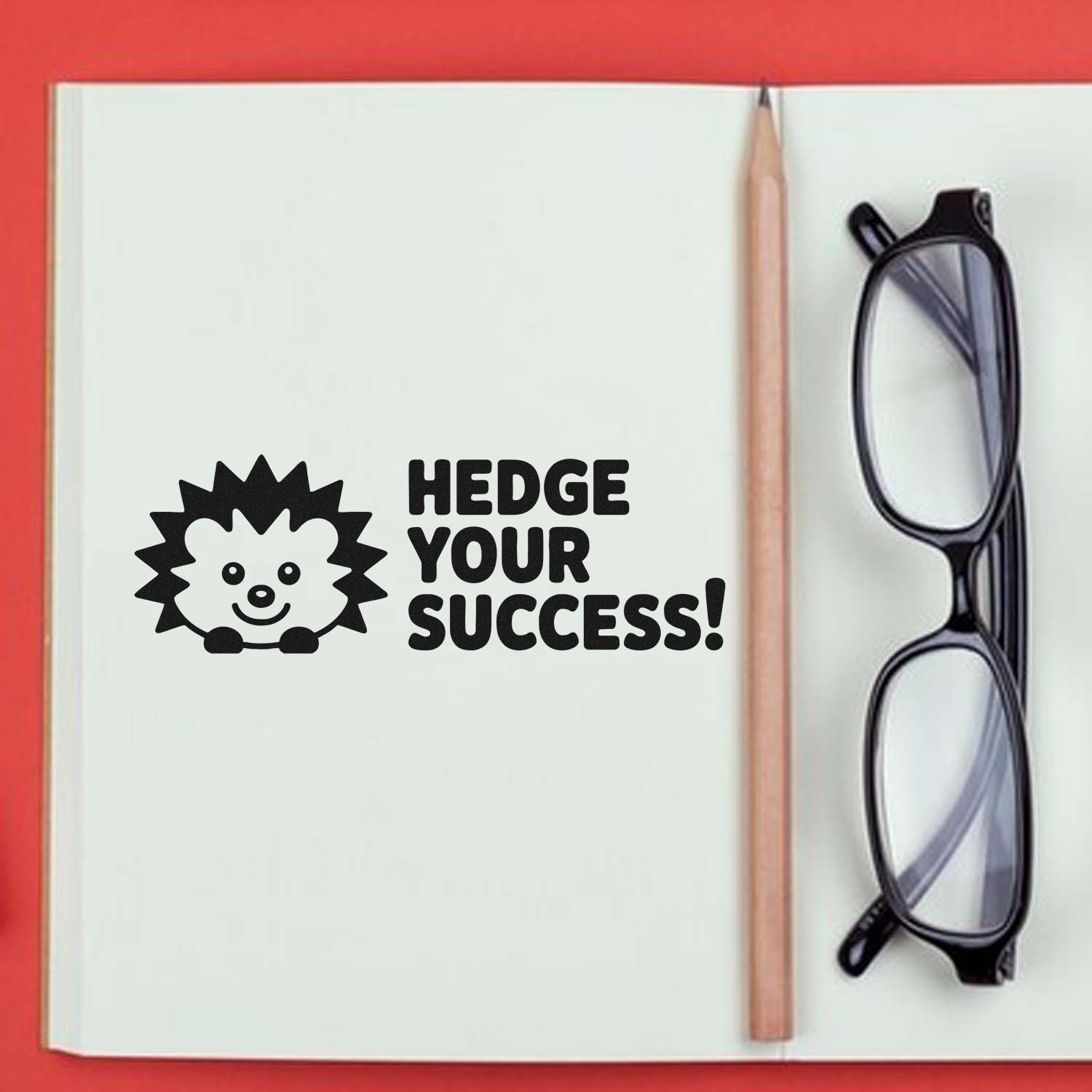 Black hedgehog stamp seal with HEDGE YOUR SUCCESS! on a white notebook, beside a pencil and black glasses on red background.