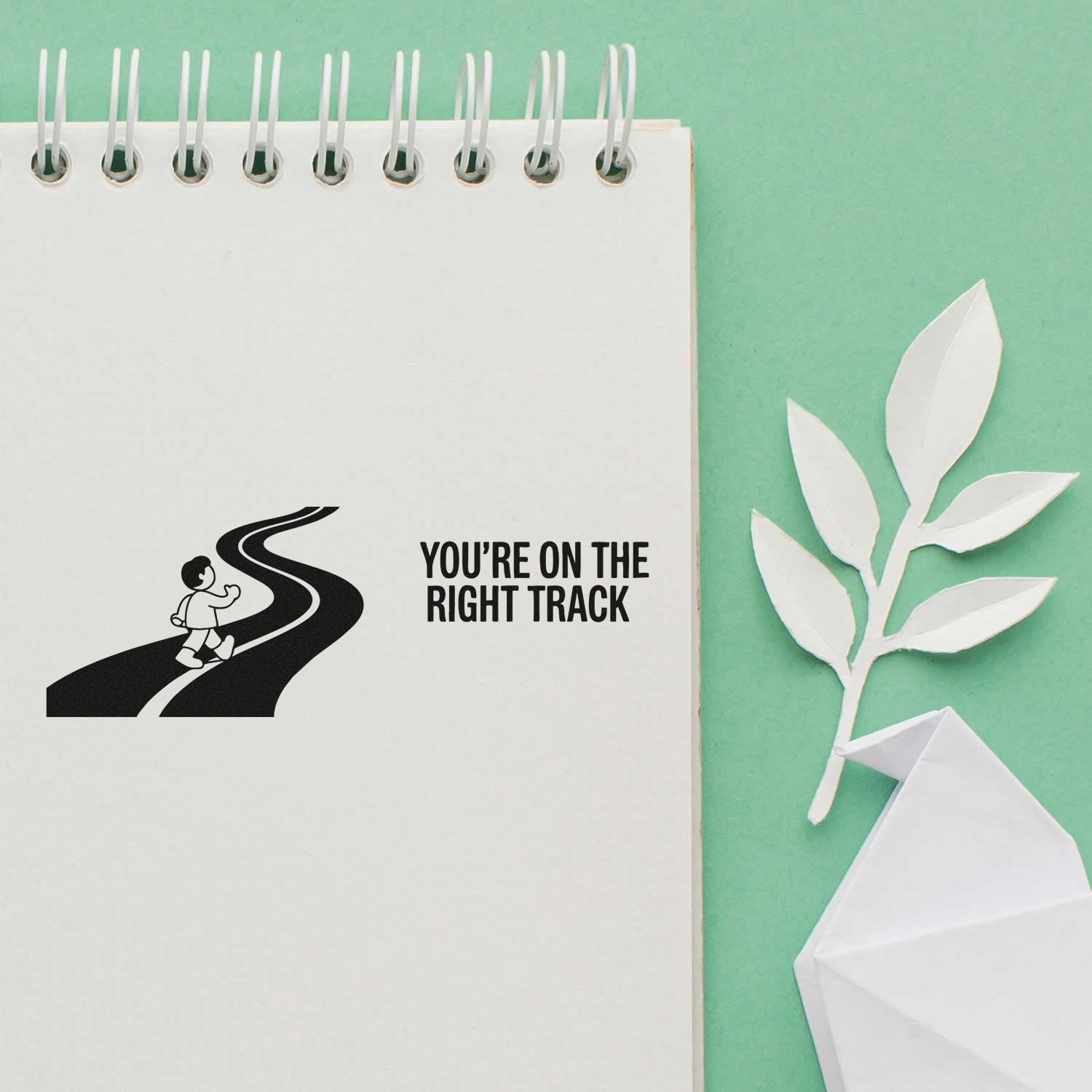 Black motivational stamp on white spiral notebook, with white paper leaf seal on mint green background.