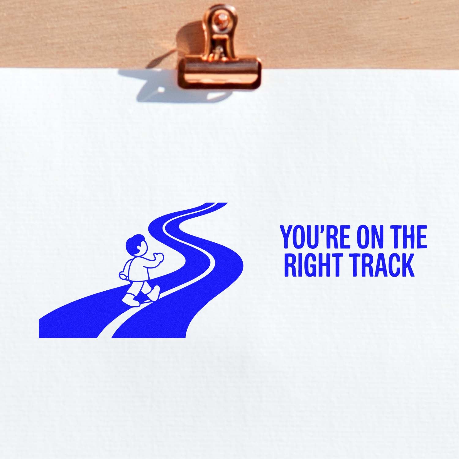 Copper clip holding white paper with blue stamp seal of a walking child on a winding road, reading Youre on the right track.