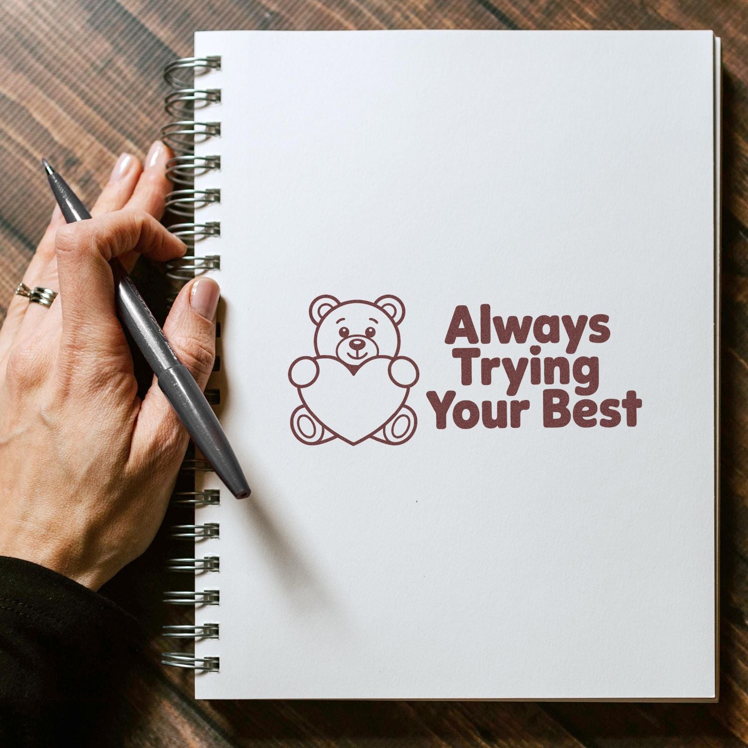 Brown teddy bear stamp/seal holding a heart with Always Trying Your Best on a white spiral notebook.
