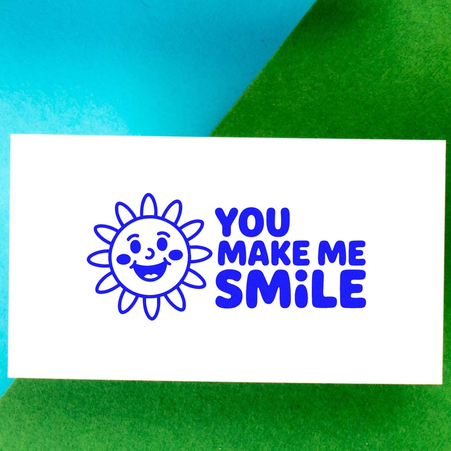 Blue stamp seal with smiling sun and YOU MAKE ME SMILE on a white card over turquoise and green background.