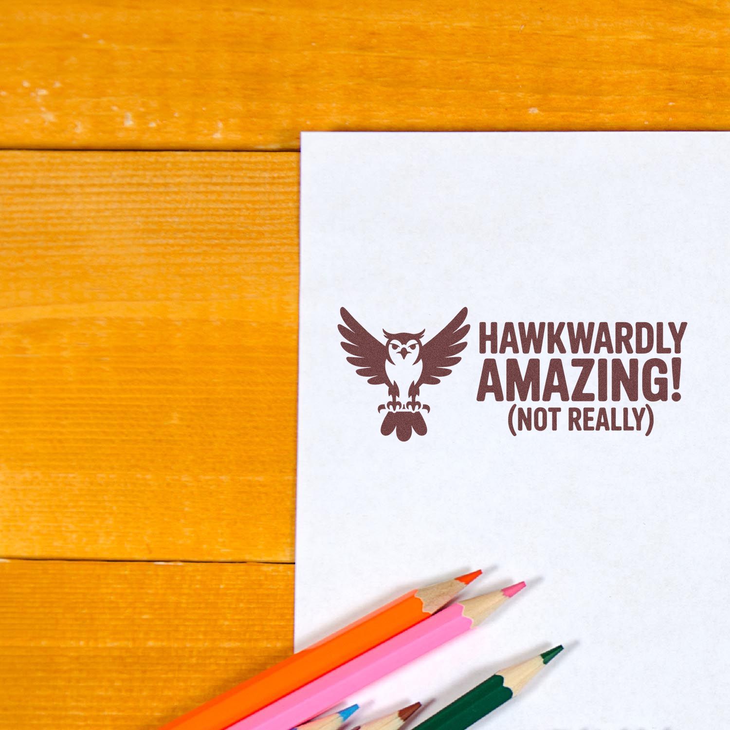 Brown stamp seal with owl and Hawkwardly Amazing! (Not Really) on white paper, with colorful pencils on orange wood.