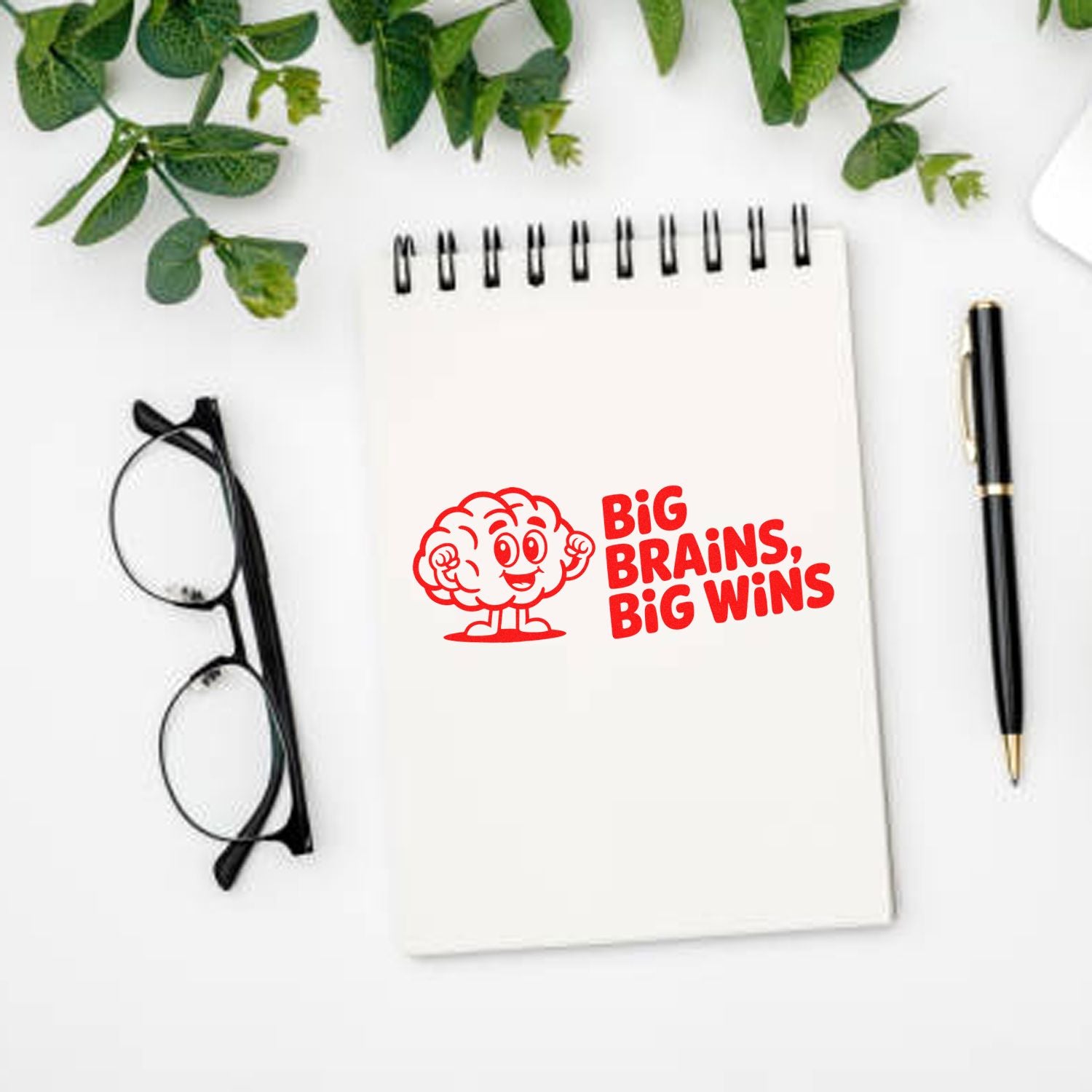 Red brain stamp and seal with Big Brains, Big Wins on a white spiral notebook, beside black pen, glasses, and green leaves.