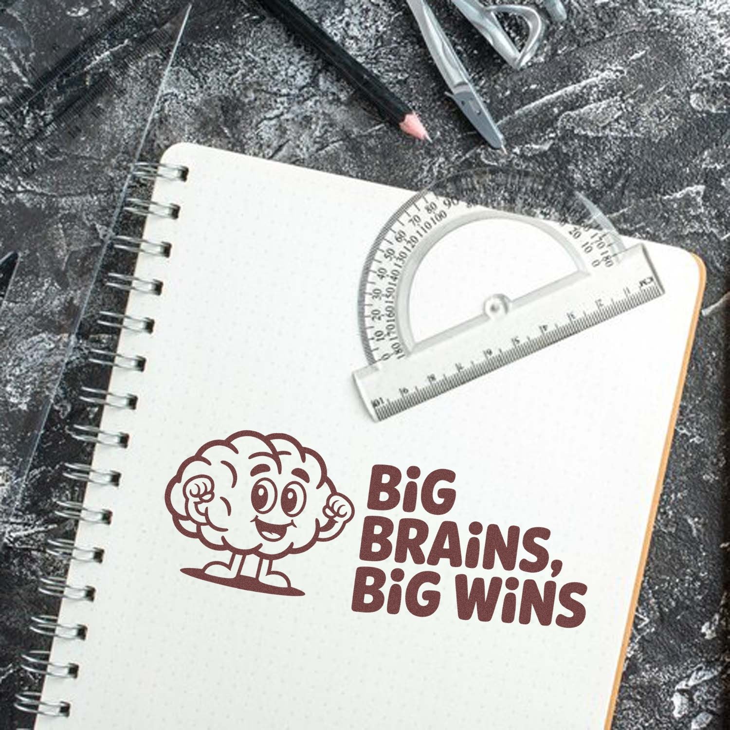 Brown brain stamp and seal design reading Big Brains, Big Wins on a white spiral notebook with clear ruler and protractor.